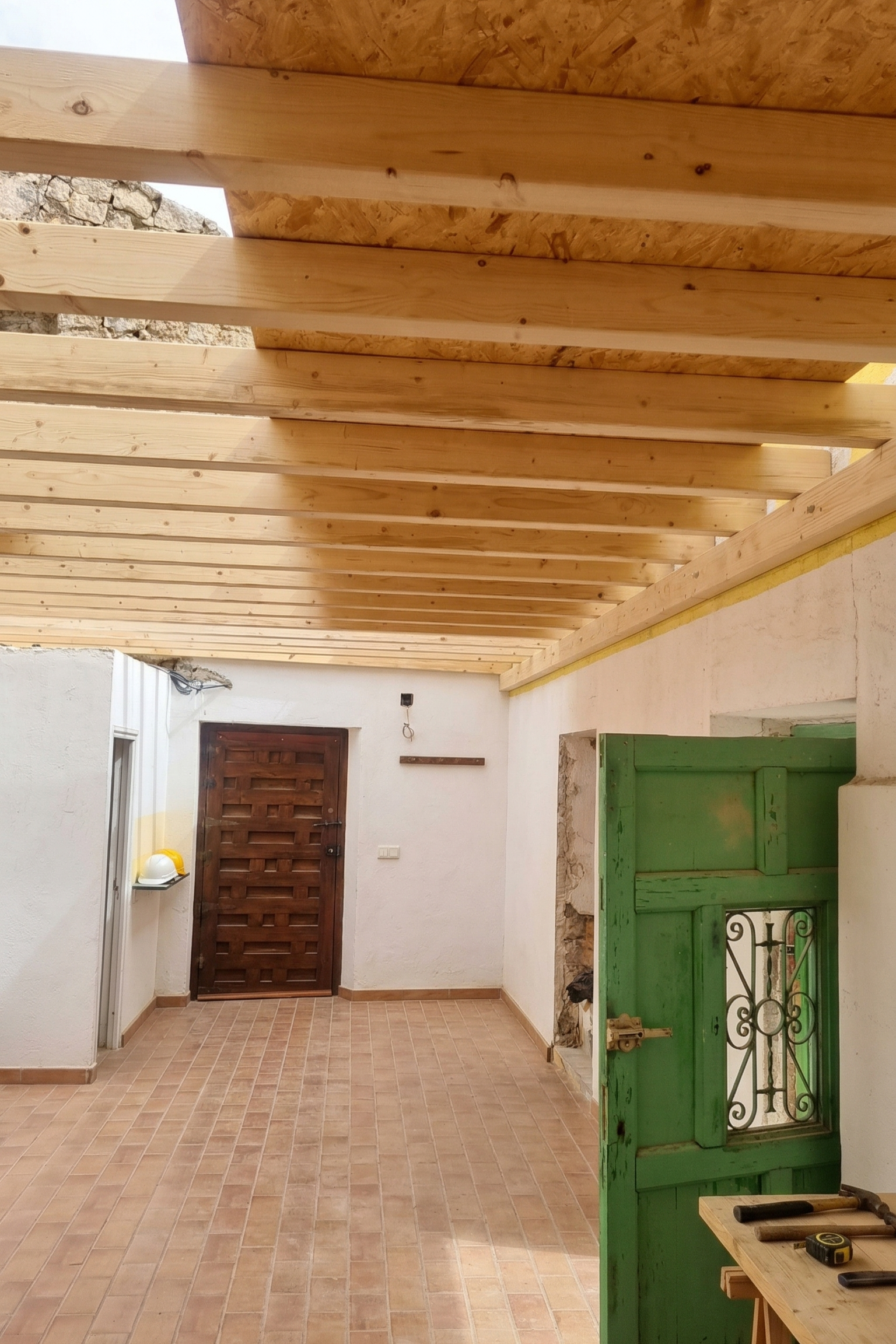 Construction of a wooden roof over a patio area with a tiled floor, a brown wooden door, a green wooden window shutter, and construction tools on a work table.