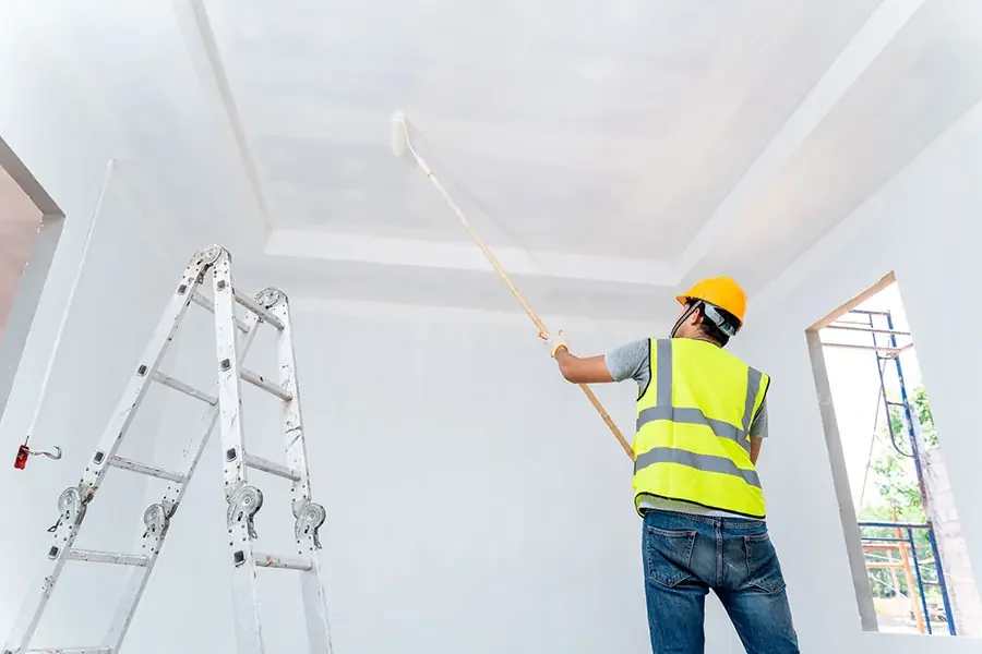 A construction worker wearing a yellow safety vest and hard hat disinfects the ceiling with a long-handled tool inside a building under construction.