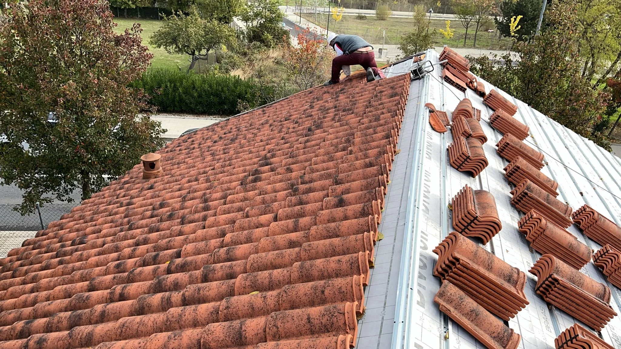 A worker installing orange ceramic roof tiles on a sloped roof, with some tiles stacked nearby. The worker is wearing a hat and bent over, working near the ridge. The background shows trees, a fence, and a road.