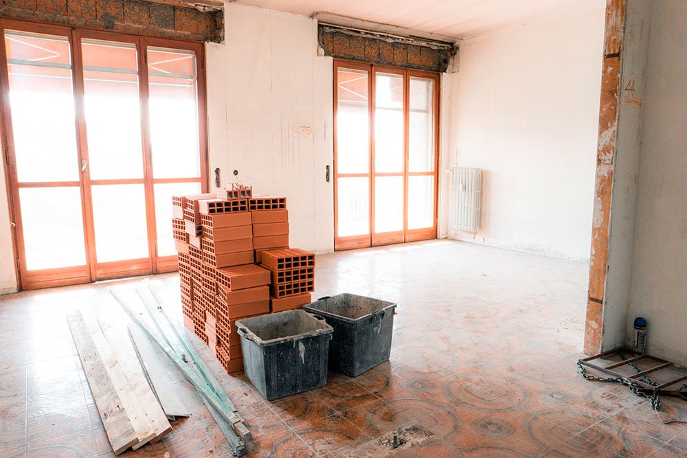 Interior of a room under construction with large windows, a pile of red bricks, two black containers, and construction tools on the floor.