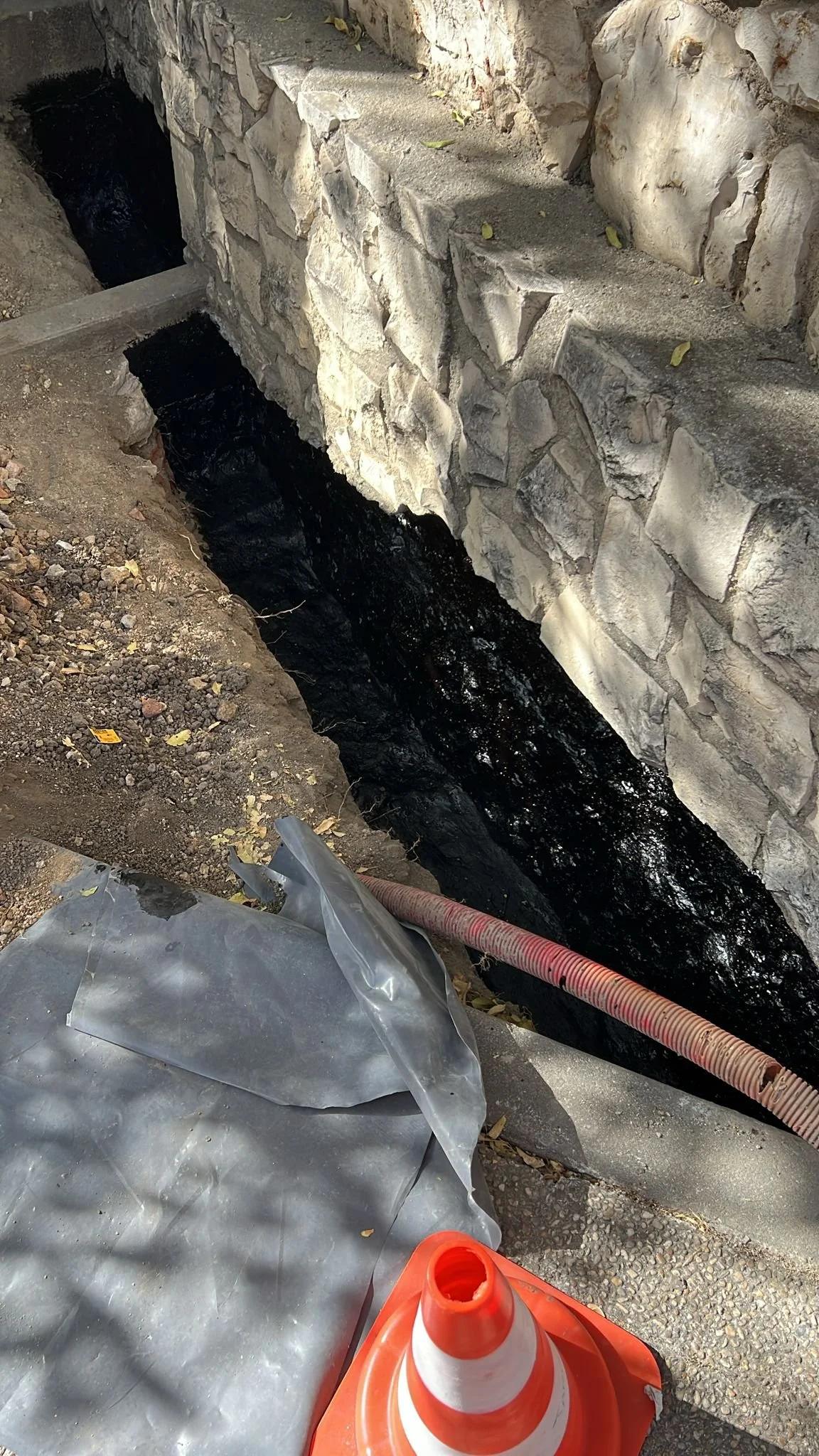A construction site showing a trench with a black pipe deeper inside, surrounded by a stone wall on one side and dirt on the other. An orange and white traffic cone and a plastic sheet are in the foreground.