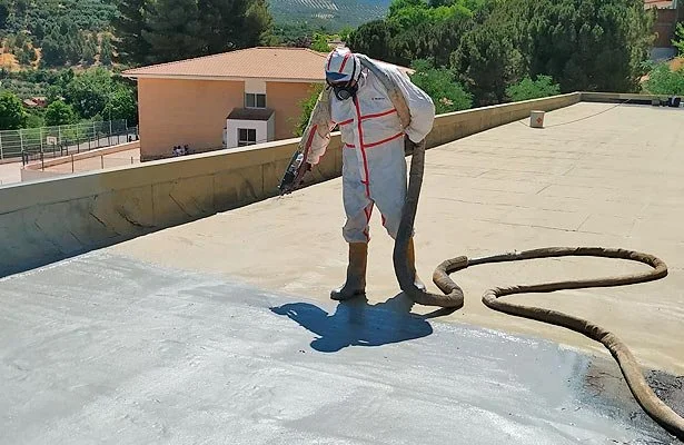 A worker on a rooftop wearing protective gear and spraying a coating, with a hose in hand and a blower on their back.