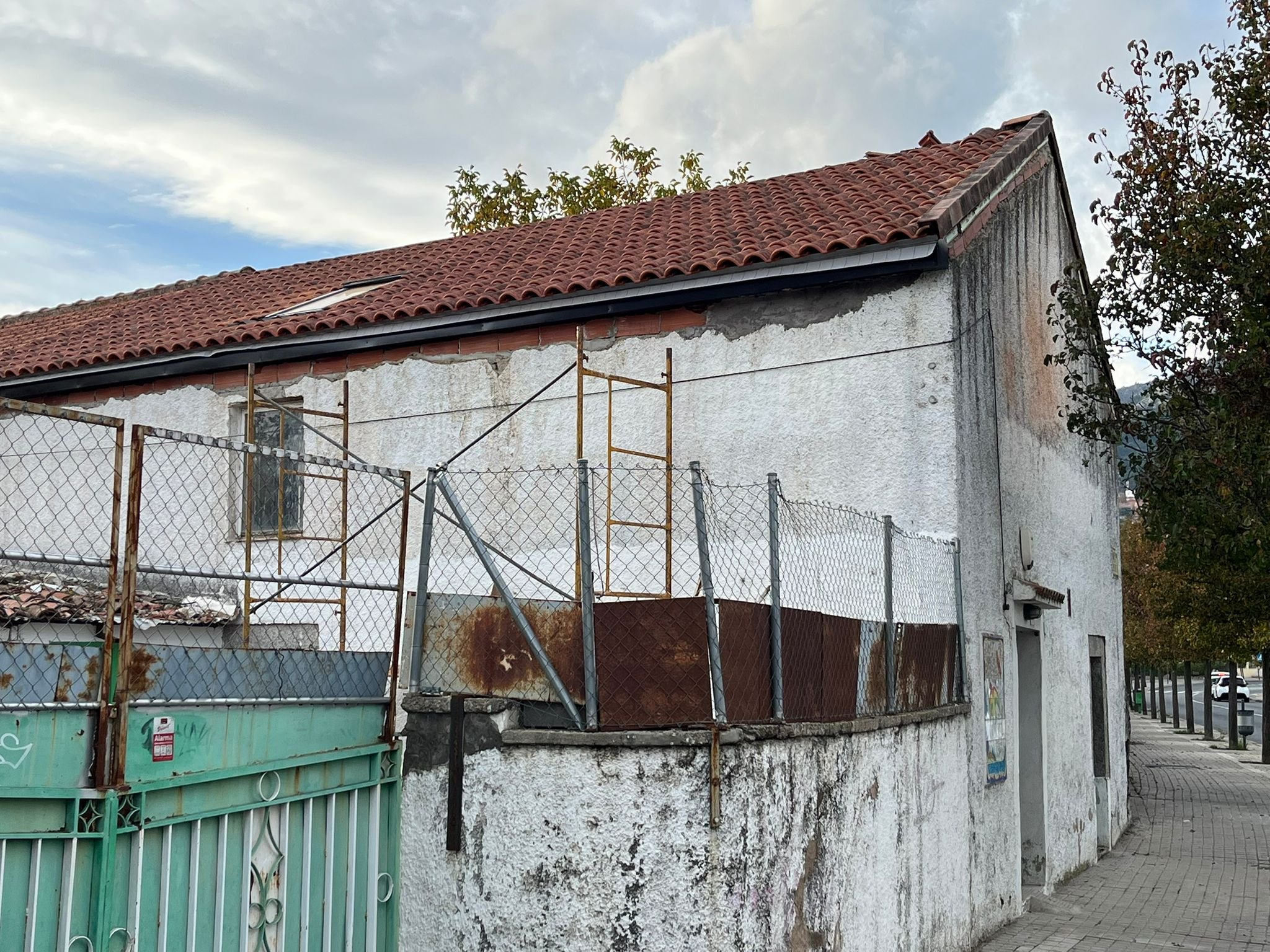 A weathered white house with a red tiled roof, chain-link fence, and a sidewalk along the right side. The house has patches of rust and peeling paint, with a small window on the side and a door near the corner.