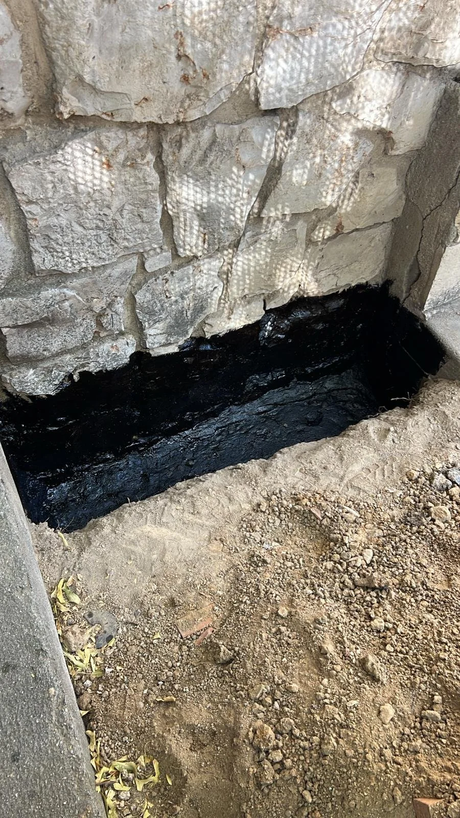 A stone wall with a rectangular hole at the bottom filled with black asphalt. The ground in front of the hole is dirt with small rocks and some fallen leaves.