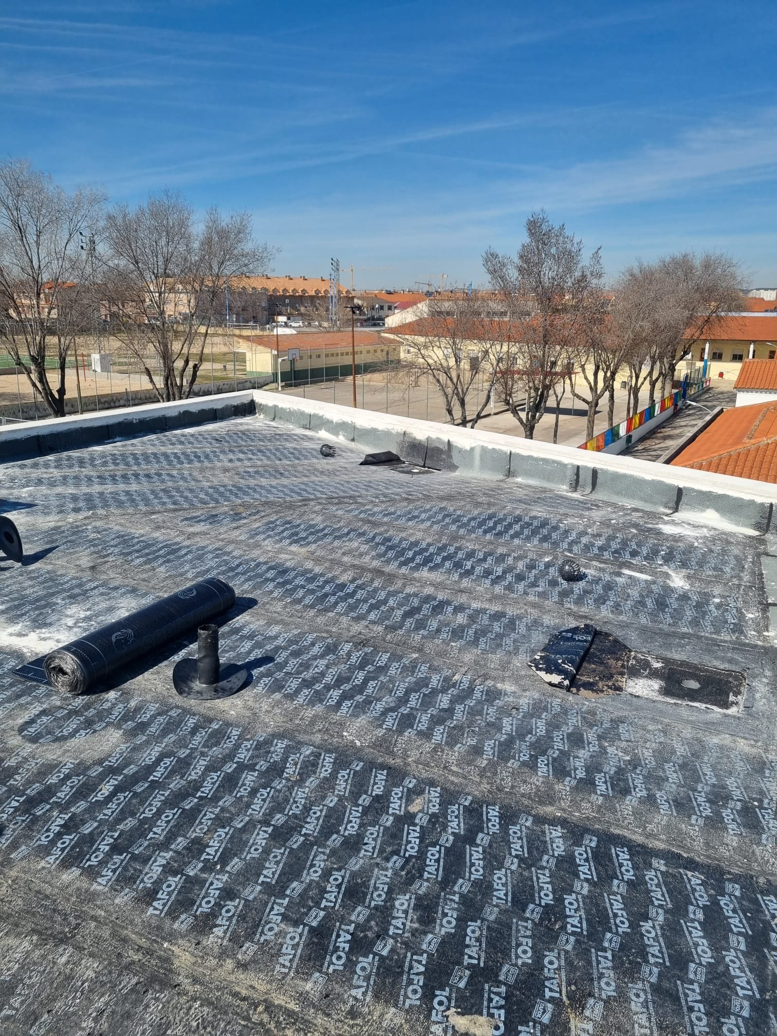 Rooftop with roofing underlayment material, vents, and debris, overlooking a schoolyard with trees, basketball courts, and a clear blue sky.