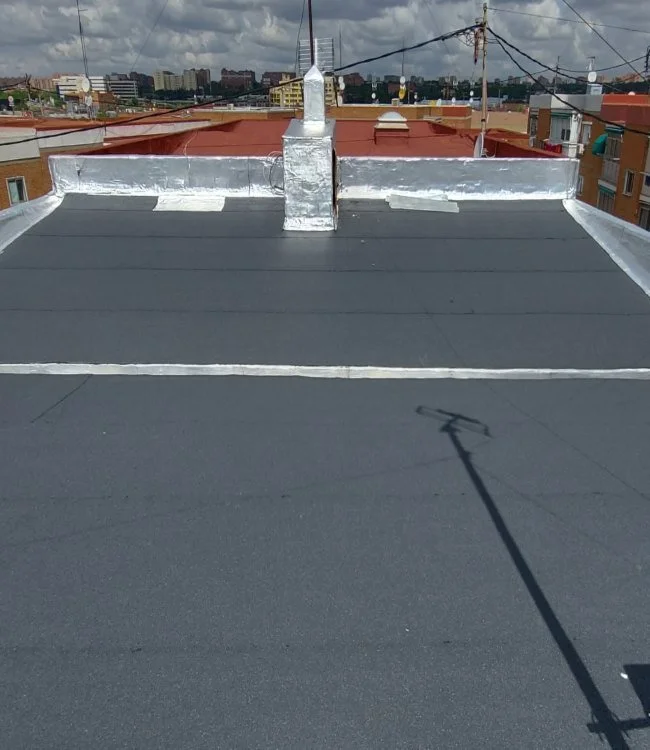 View of a flat rooftop with a black surface, silver flashing around the edges and chimney, and city buildings in the background.
