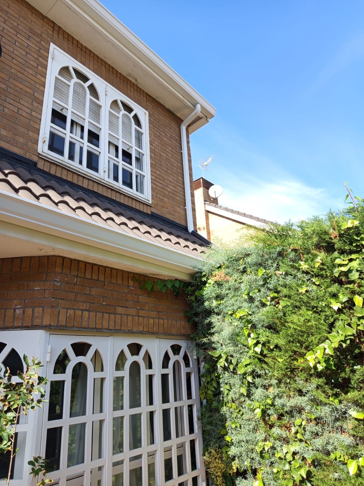 The exterior of a brick house with white decorative window frames, a sloped tiled roof, and greenery covering part of the wall. The house has two stories with a window on each level, and the sky is clear and blue.