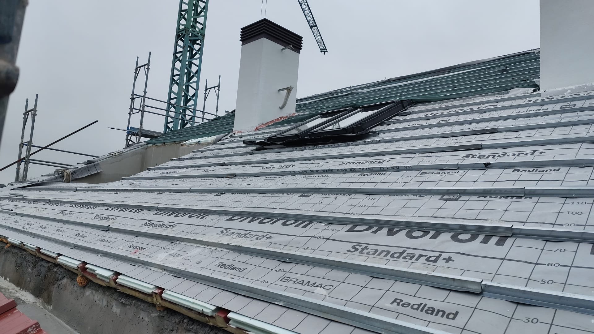 Roof under construction with underlayment, metal flashing, and a nearby chimney, with scaffolding and a crane in the background.