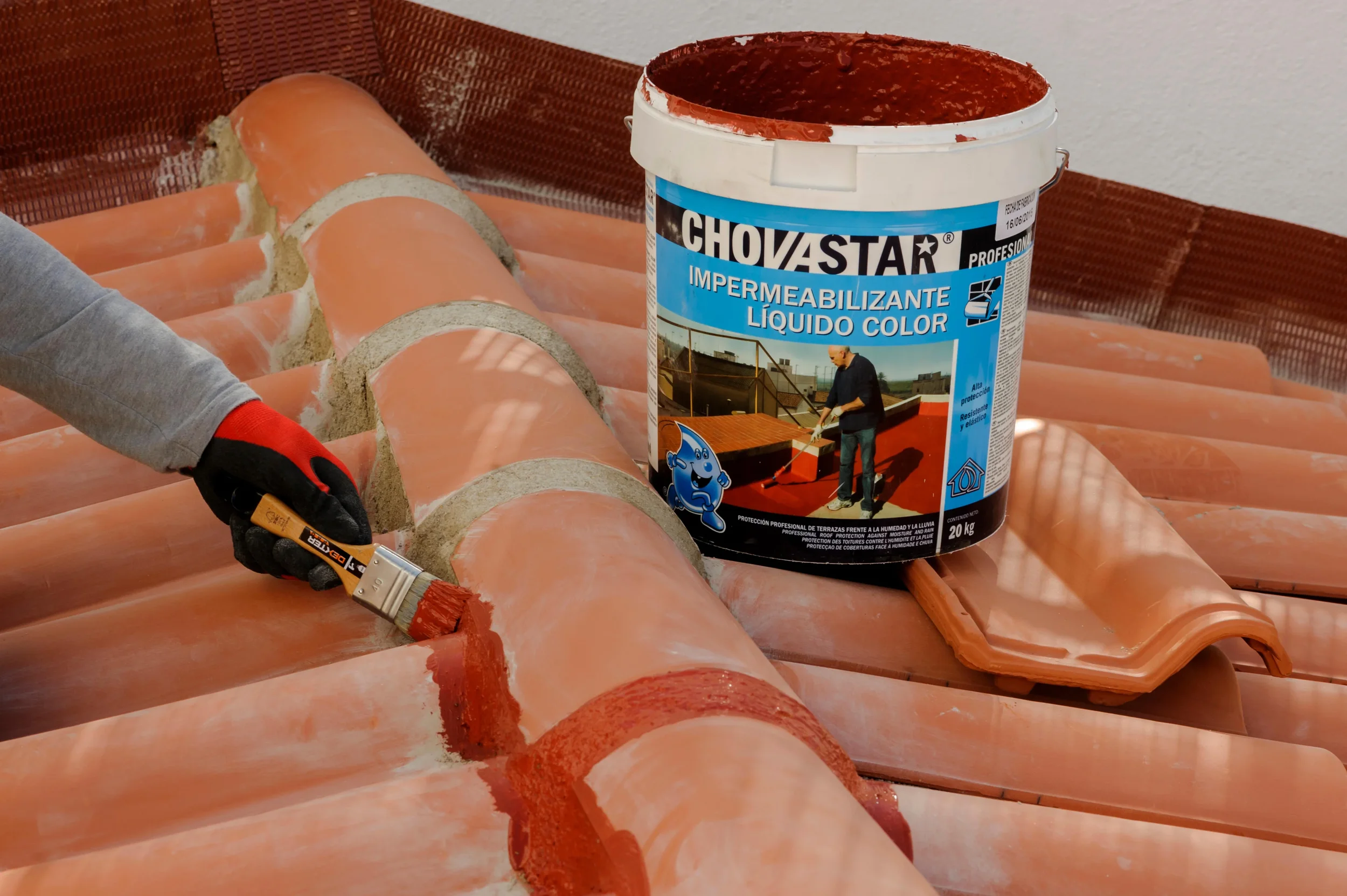 A person applying red waterproofing liquid to clay roof tiles with a small paintbrush, with a large bucket of waterproofing liquid in the background.