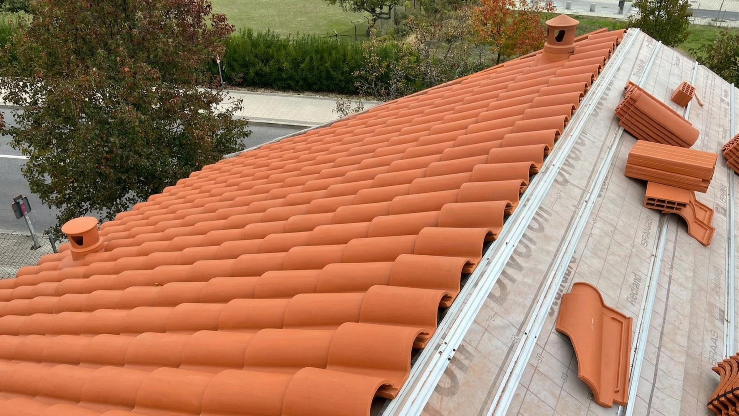 A roof under construction with orange clay tiles, metal roof flashing, and various uninstalled tiles on a partially completed section. The background shows trees and a sidewalk.