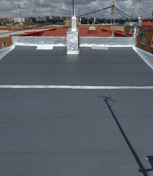 View of a rooftop with a black roofing material, silver flashing around the chimney, and shadows of utility poles and wires, with city buildings and partly cloudy sky in the background.