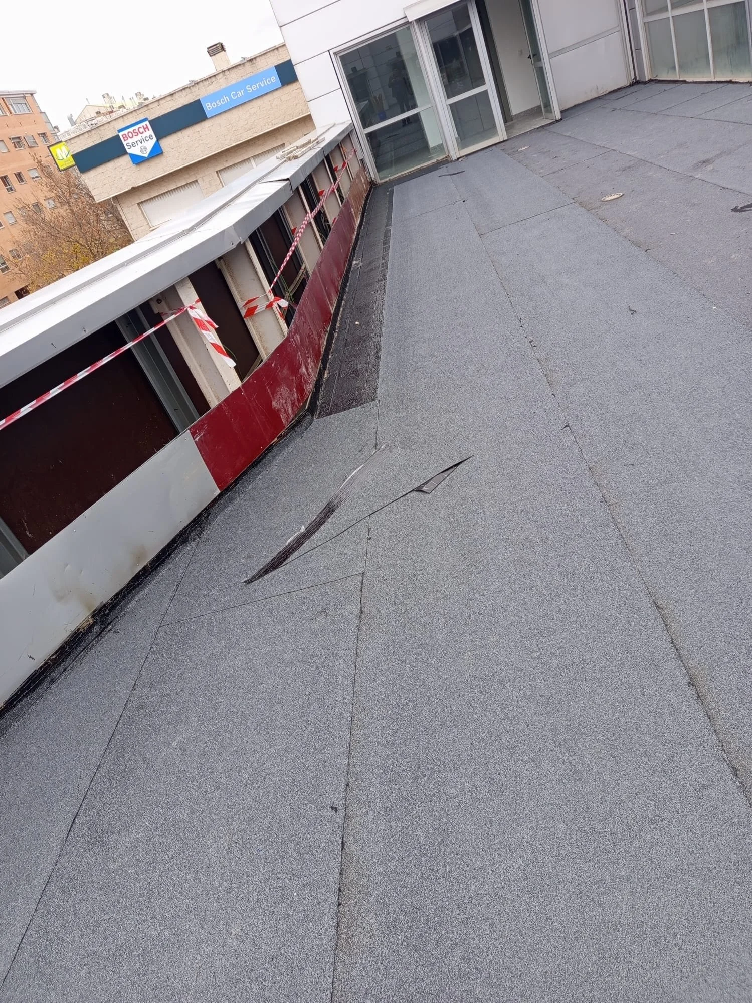 A rooftop with gray roofing material featuring a large, damaged area with torn sections. There are orange and white striped caution tapes along the edge of the roof, and a building with large glass windows and doors is visible in the background. A si