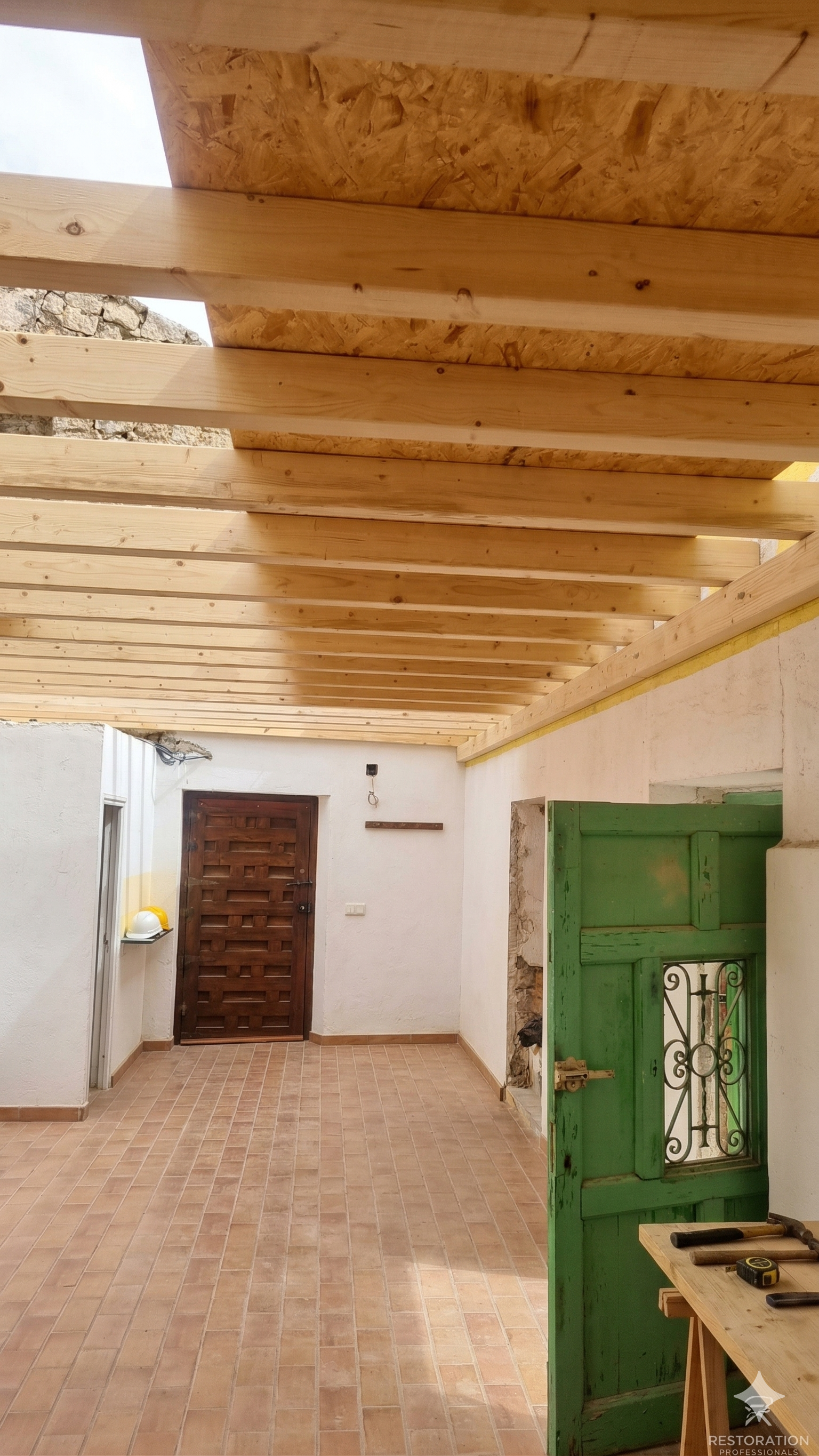 Interior of a room under construction with a wooden ceiling, tiled floor, and a green door. Tools on a work table and a brown wooden door at the back.