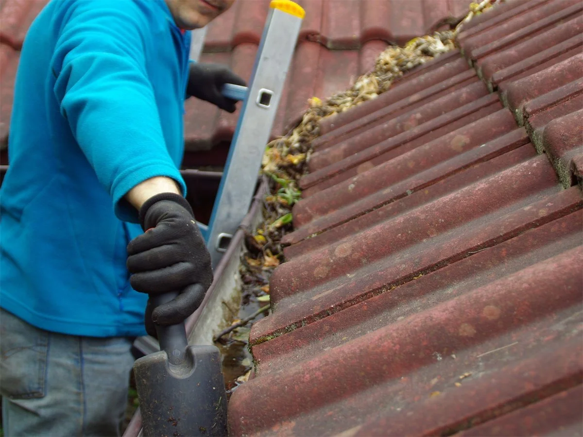 A person in a blue jacket and black gloves using a trowel to clean leaves and debris on a red tiled roof.