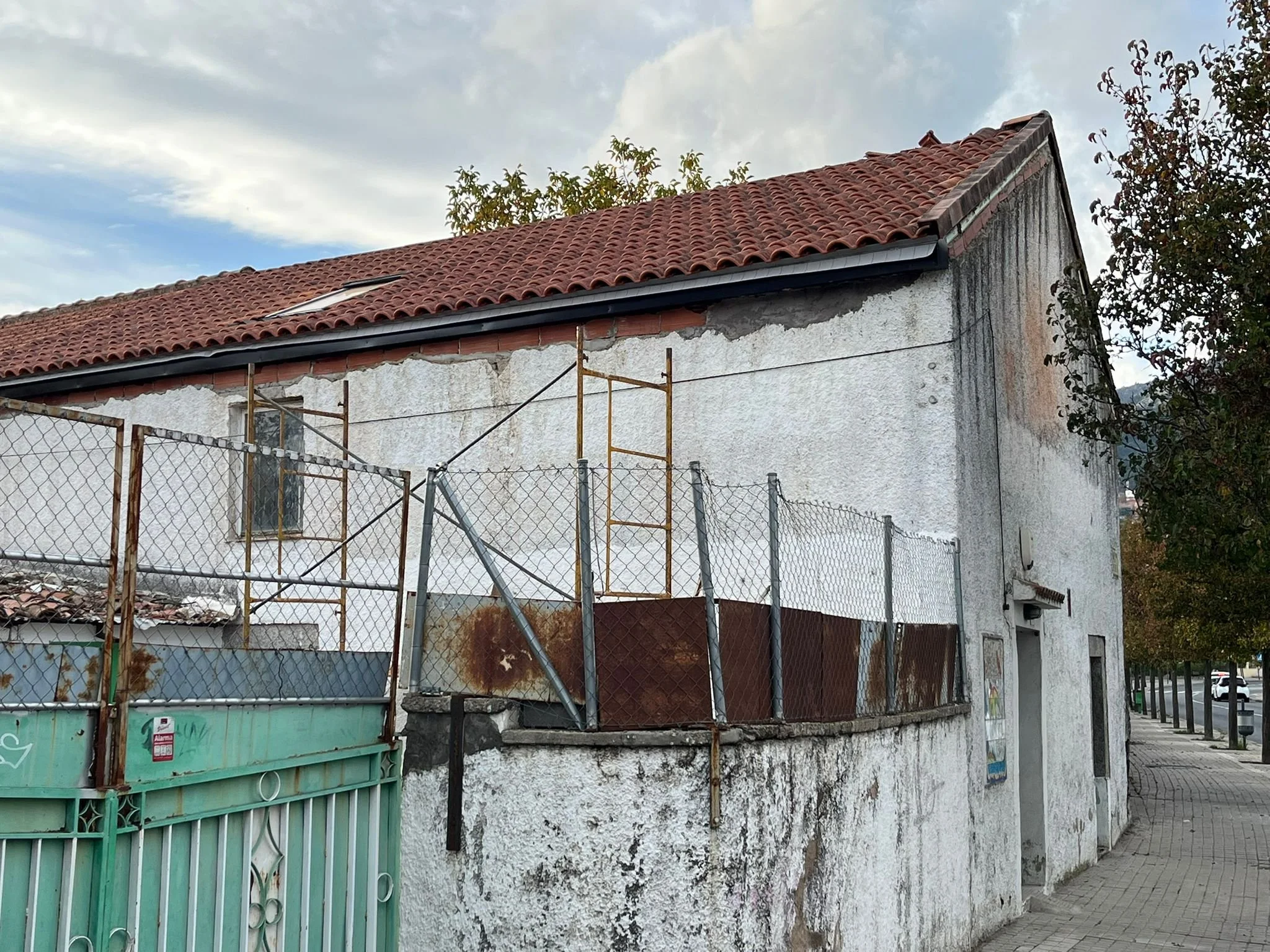 A two-story building with a red tile roof, white stucco walls, and a rusted chain-link fence in front. There is a small window on the left side and a larger door on the right side. The building shows signs of wear and weathering.