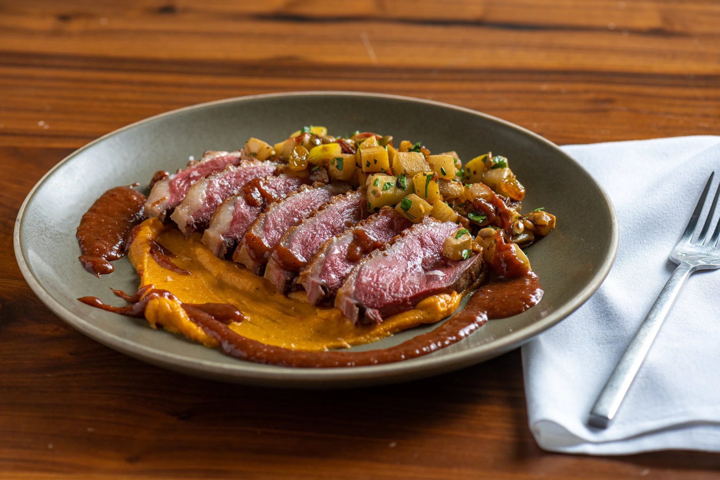 Serrano pepper sauce, medium rare sliced beef with sauce, on a bed of squash purée, topped with diced vegetables, on a grey plate, set on a wooden table with white napkin and fork.