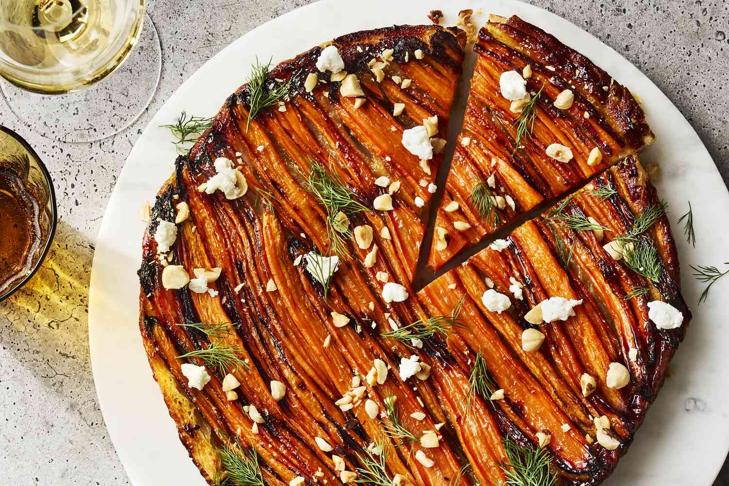 A carrot tart with roasted carrots drizzled with a glaze, topped with chopped nuts and fresh herbs on a white plate.