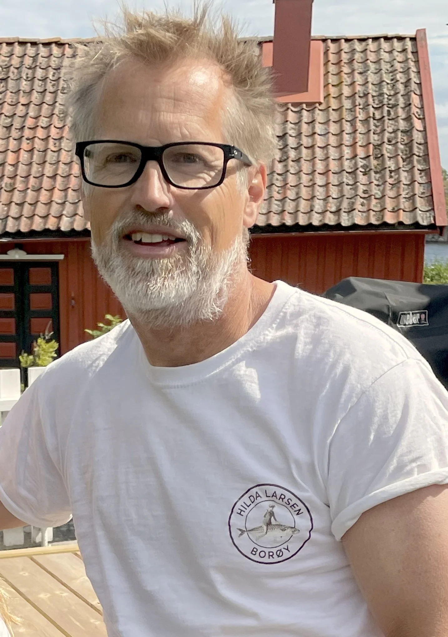 A man with glasses and a white beard wearing a white T-shirt with a logo, outdoors in front of a red house with a tiled roof.
