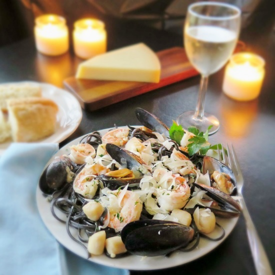 Seafood pasta with mussels, shrimp, and herbs on a white plate, accompanied by a glass of white wine and candles in the background.