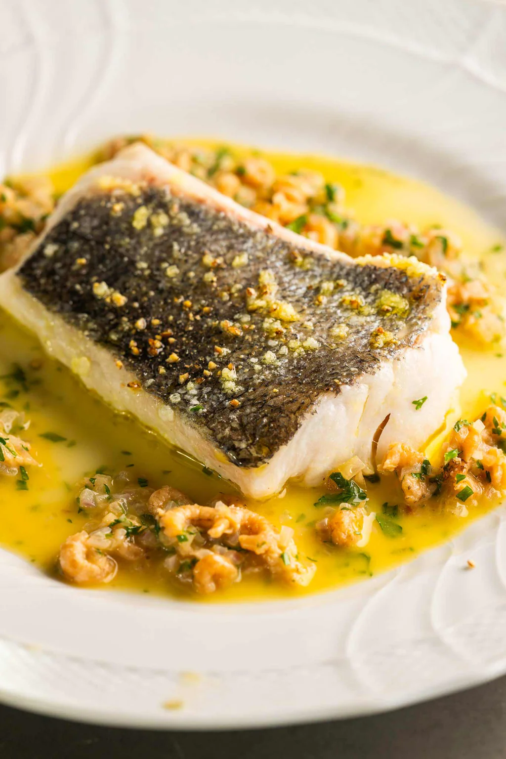 A cooked fish fillet with crispy skin on a white plate, topped with herbs and served with a lemon butter sauce and chopped nuts.