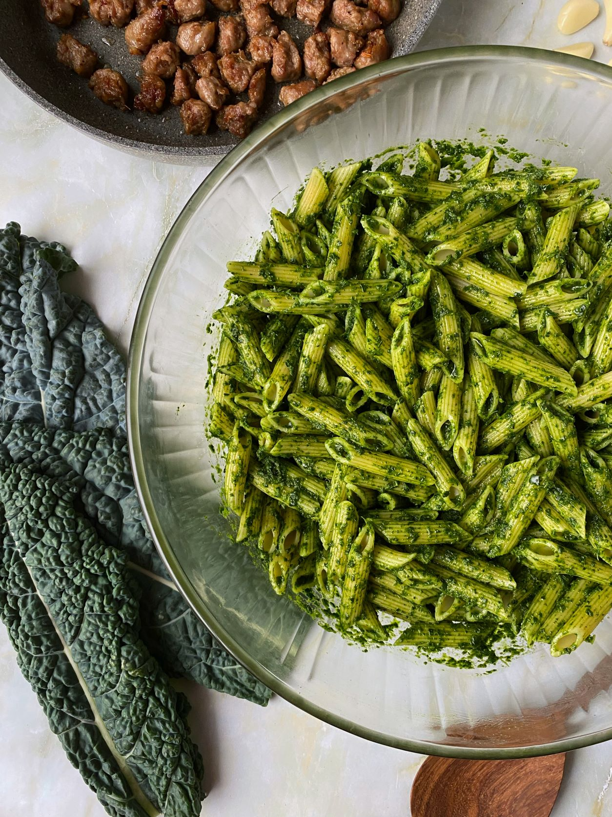 A glass bowl of cooked pesto pasta with kale leaves and a skillet of cooked ground beef in the background.