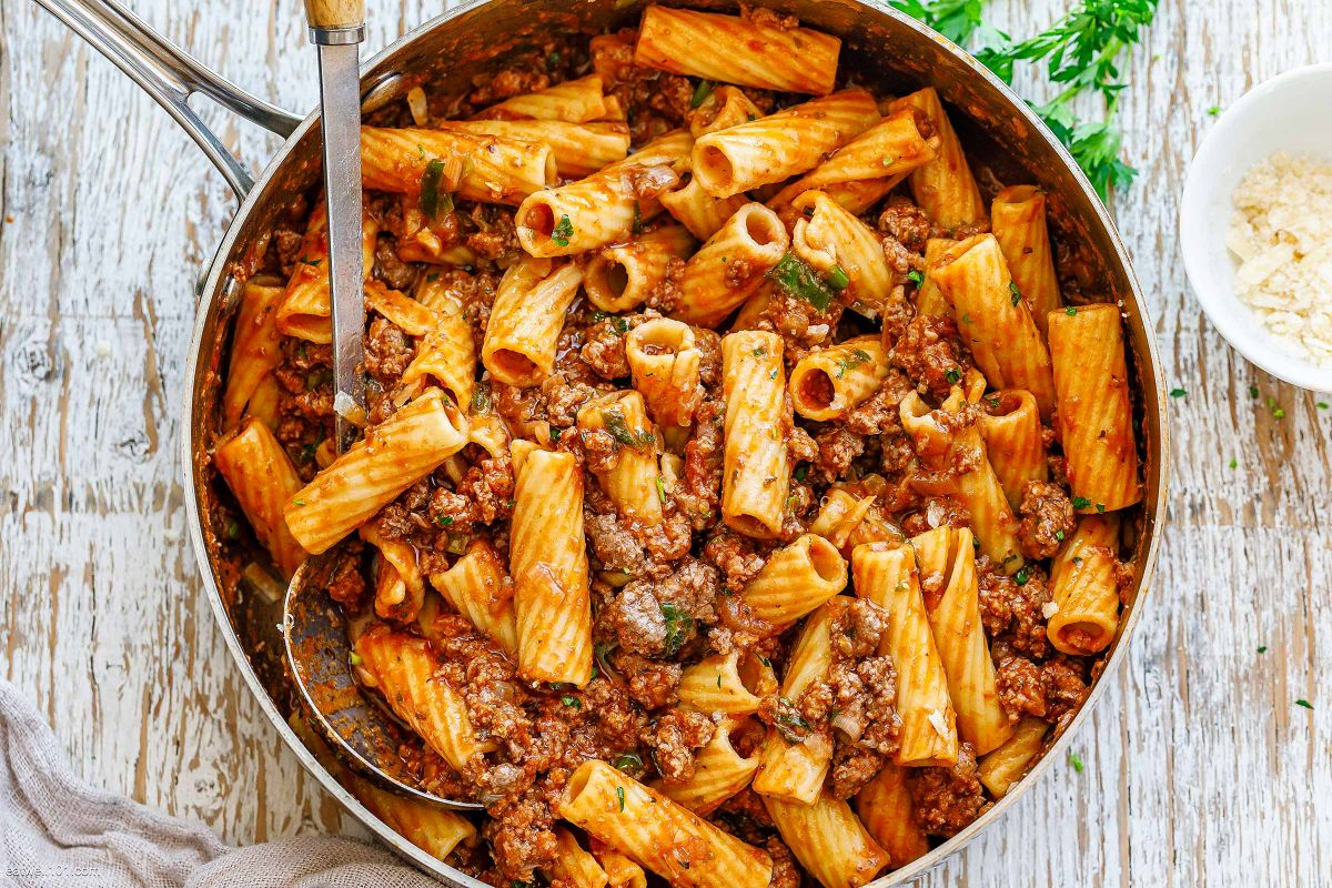 A skillet of cooked rigatoni pasta with ground beef and tomato sauce, garnished with chopped herbs. A spoon rests in the skillet, and a small bowl of grated cheese is nearby on a rustic wooden surface.