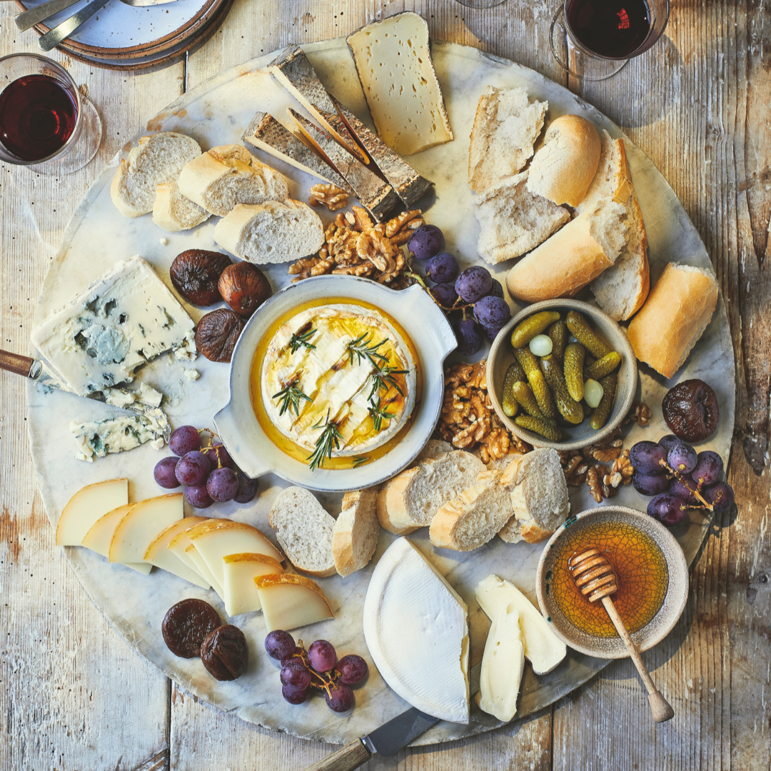 A round cheese platter with various cheeses, bread, grapes, figs, walnuts, and accompaniments like honey and a creamy dip, served on a rustic wooden table with glasses of red wine.