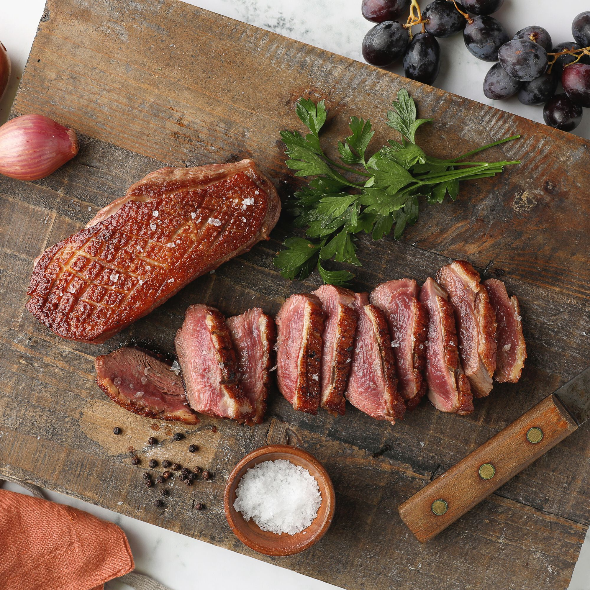 Sliced cooked steak with salt, parsley, garlic, black peppercorns, and grapes on a wooden cutting board.