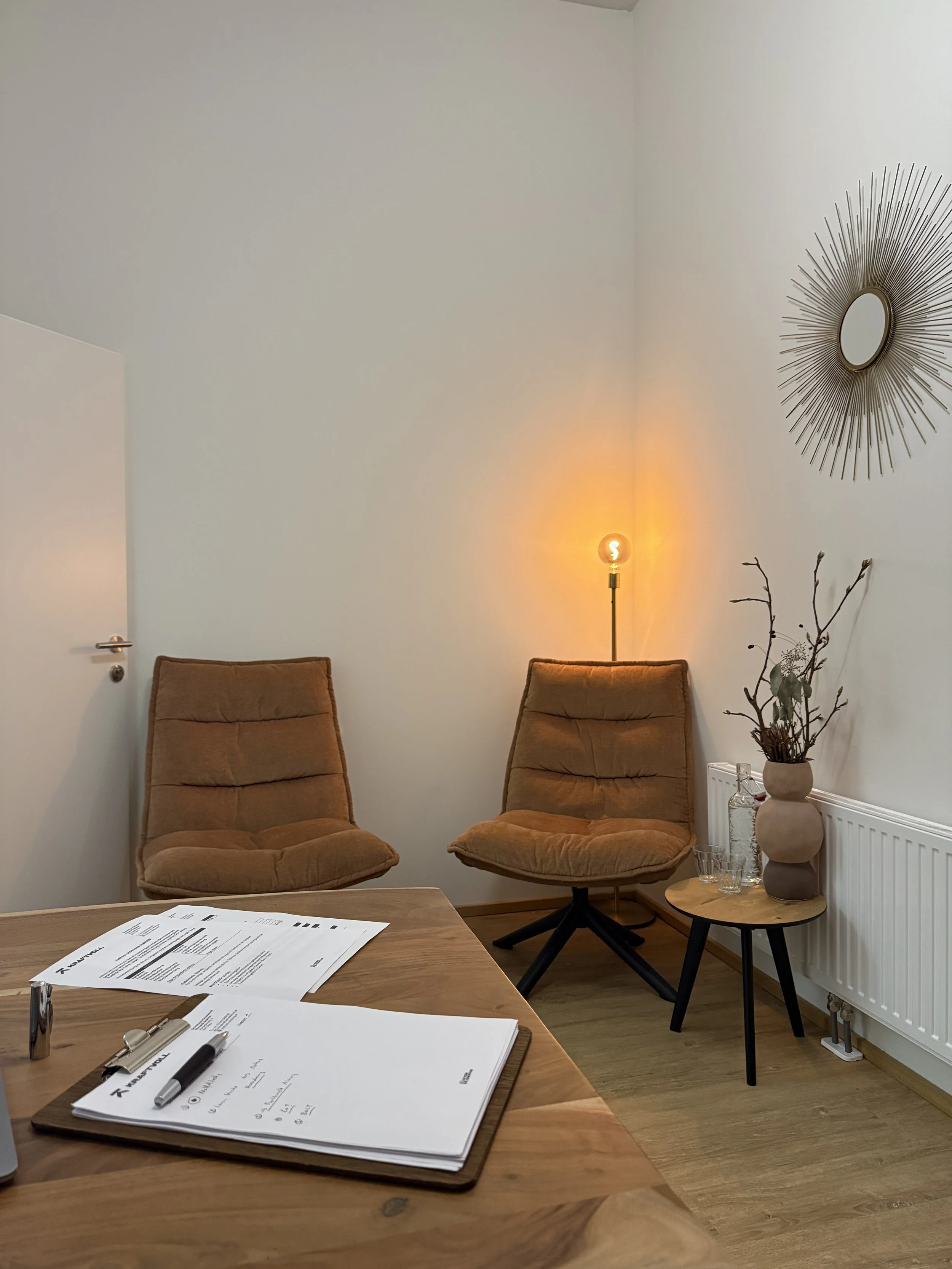 Ein Büro mit zwei braunen Sesseln, einem kleinen runden Tisch mit einer Vase und Gläsern, einem Holzschreibtisch mit Papier und Stiften, einer Stehlampe mit warmem Licht, einer Wanddekoration und einer weißen Wand.