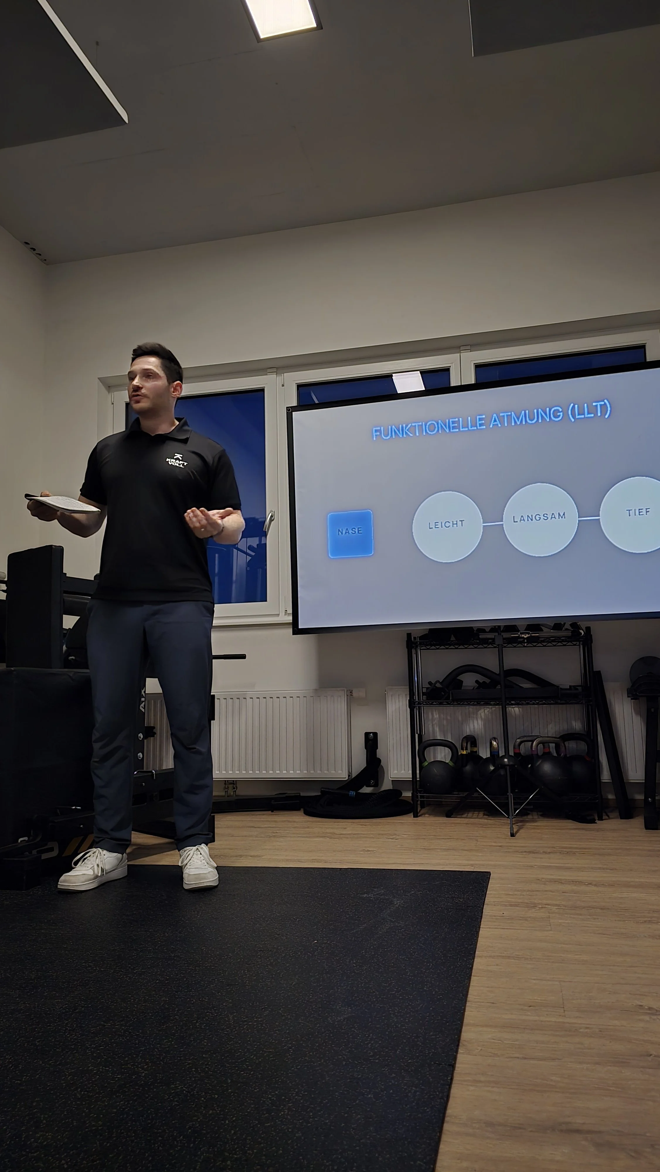 Ein Mann hält in einem Fitnessraum eine Präsentation über funktionelle Atmung, mit einem Bildschirm im Hintergrund, auf dem die verschiedenen Atemtypen in Deutsch beschrieben sind.