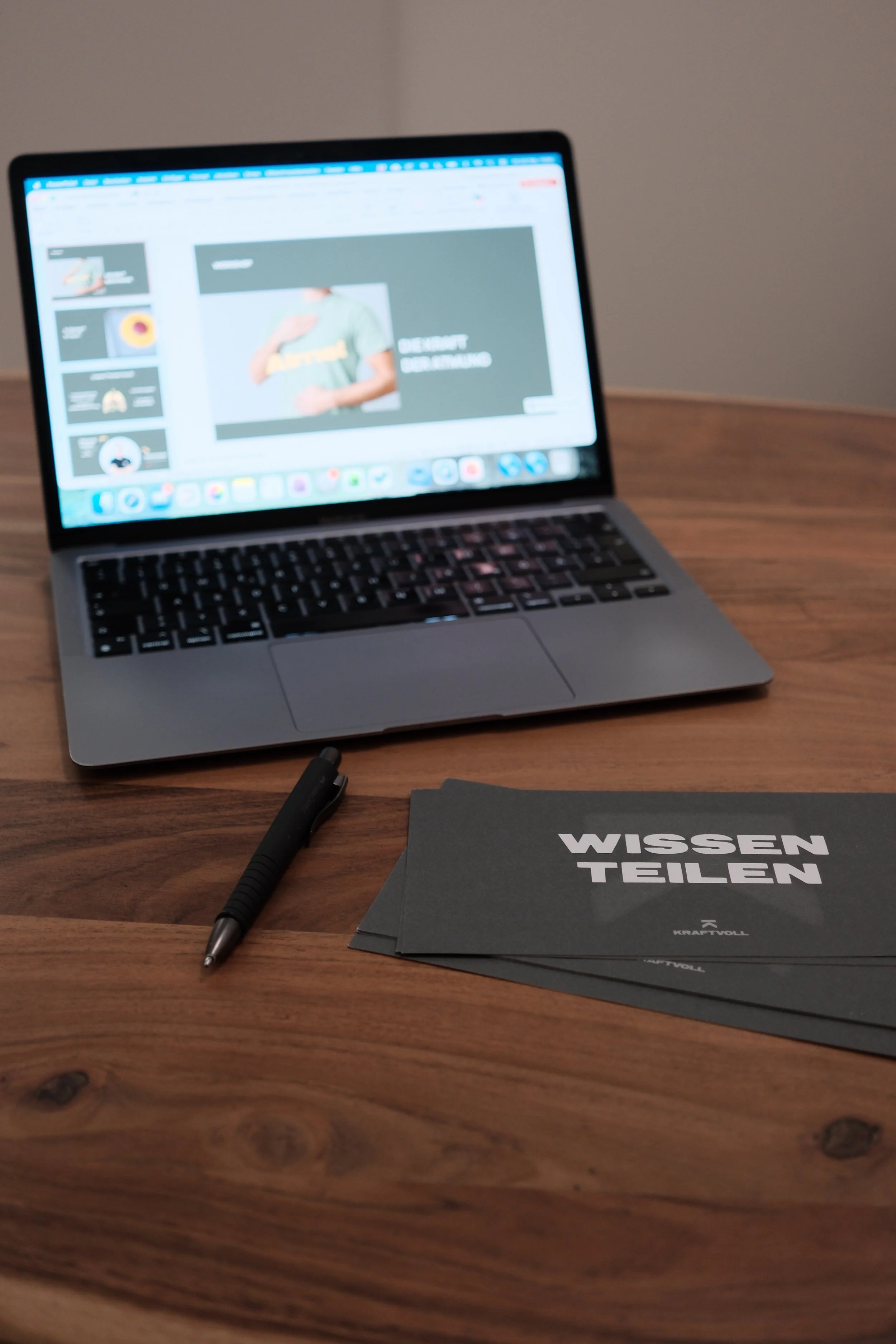 Ein Laptop, ein schwarzer Kugelschreiber und mehrere schwarze Broschüren auf einem Holztisch. Die Broschüren haben den Titel 'WISSEN TEILEN'.