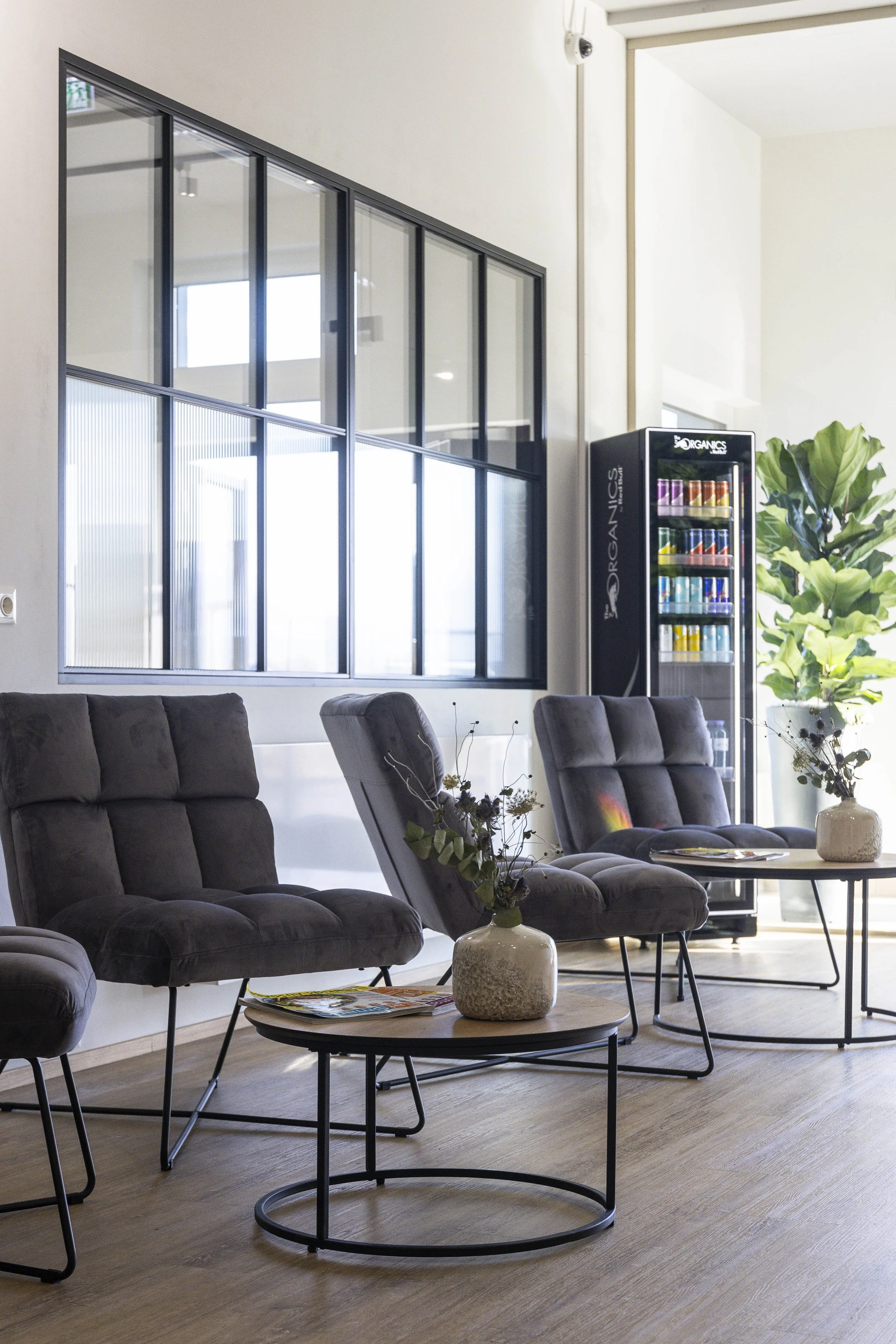 Wartebereich mit drei grauen Sesseln, niedrigen Tischen mit Dekorvasen und Zeitschriften, Pflanzen und Getränkeautomat im Hintergrund, große Fenster mit schwarzem Rahmen.