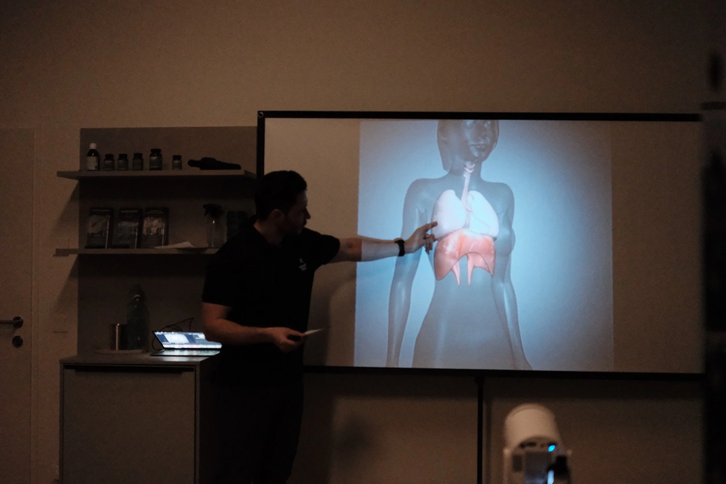 Ein Mann zeigt auf eine projizierte anatomische Darstellung eines menschlichen Herzens auf einer Leinwand, während er während einer Präsentation in einem dunklen Raum spricht.