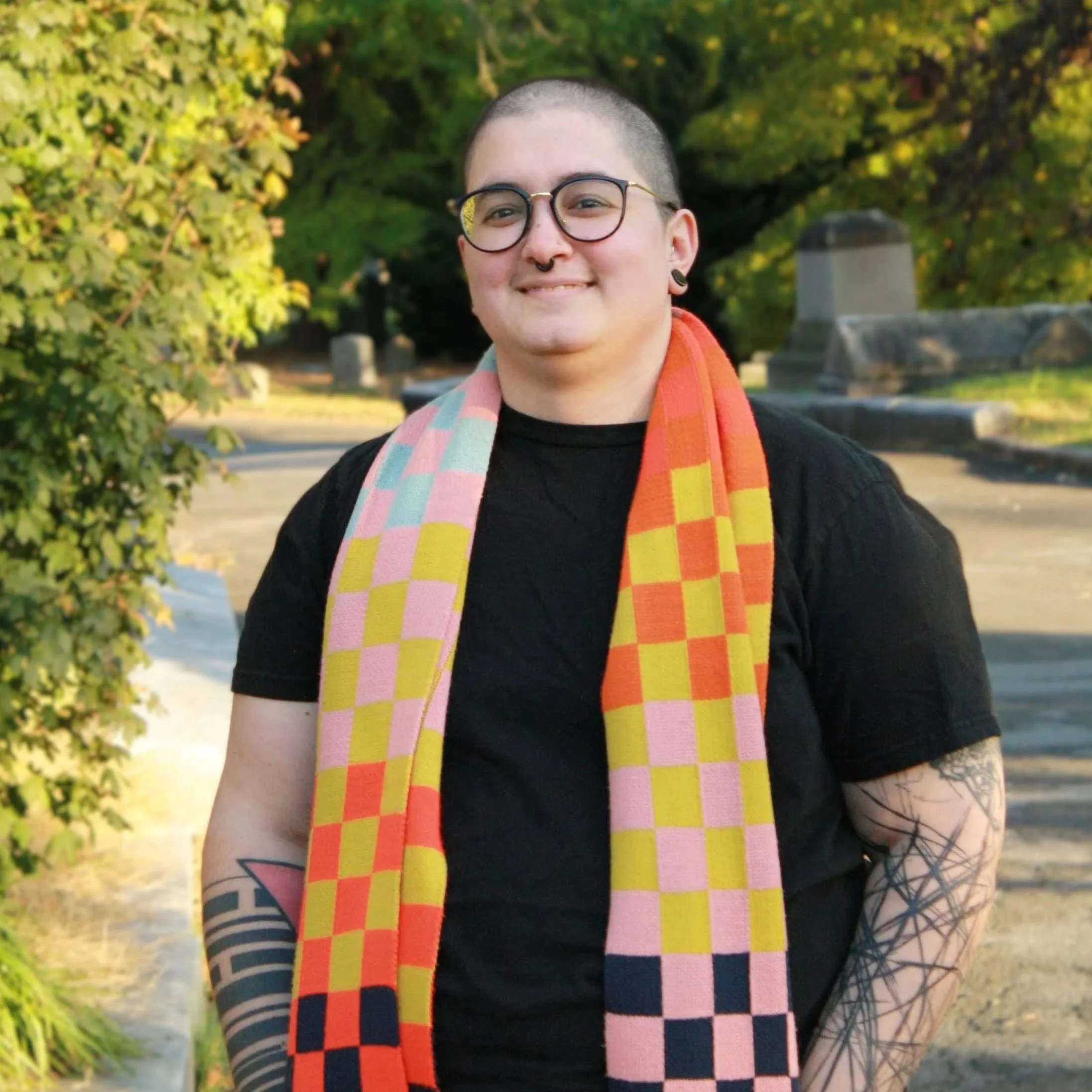 MC is standing and smiling on a leafy path. MC is a white man with glasses, facial piercings, and many tattoos.
