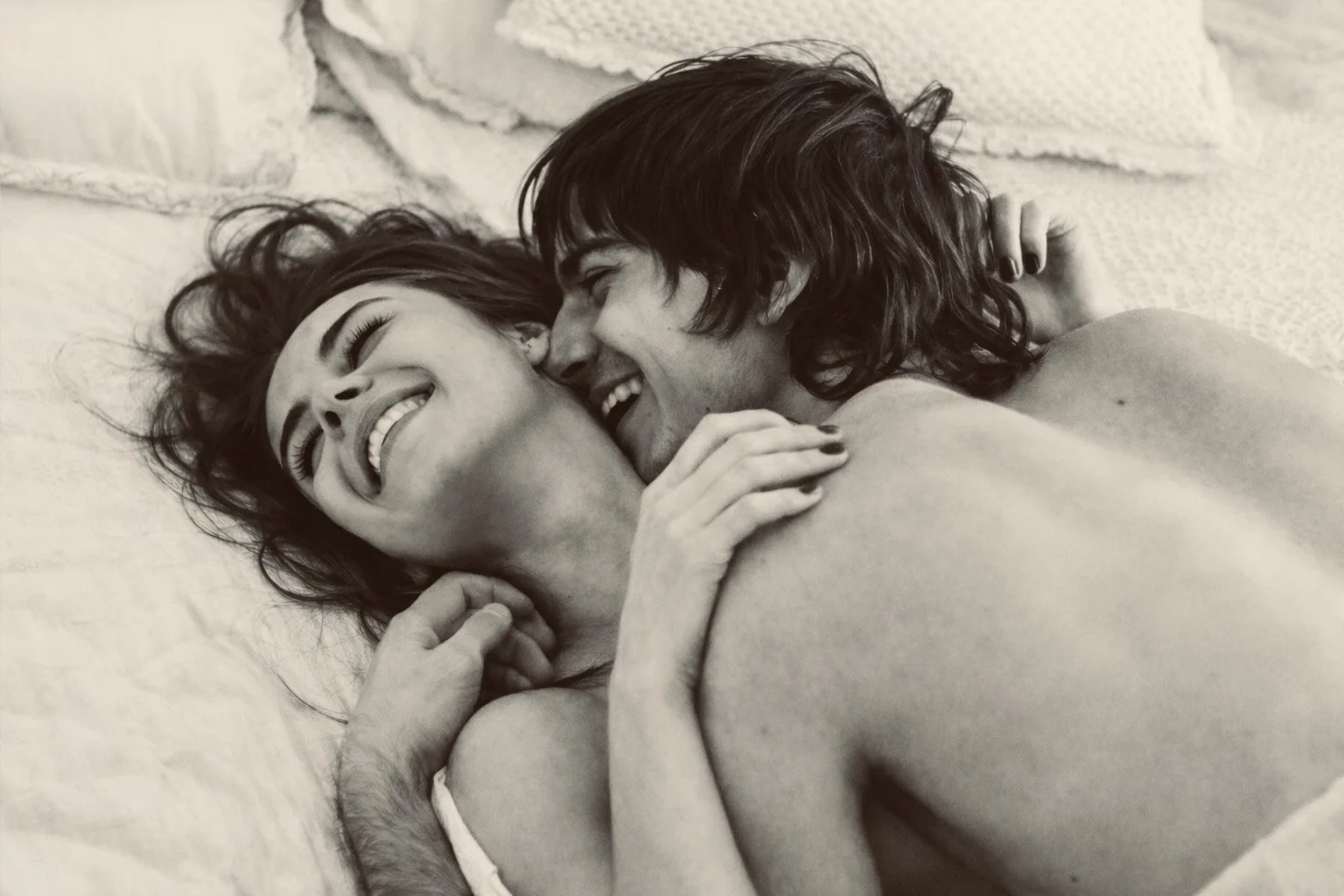 A black and white photo of a couple lying in bed, smiling and cuddling closely. The woman has short hair and is laughing, while the man has slightly longer hair and is smiling.
