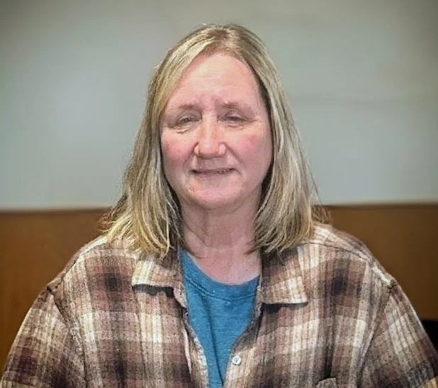 A woman with shoulder-length blonde hair, wearing a plaid shirt over a blue top, sitting against a neutral background.
