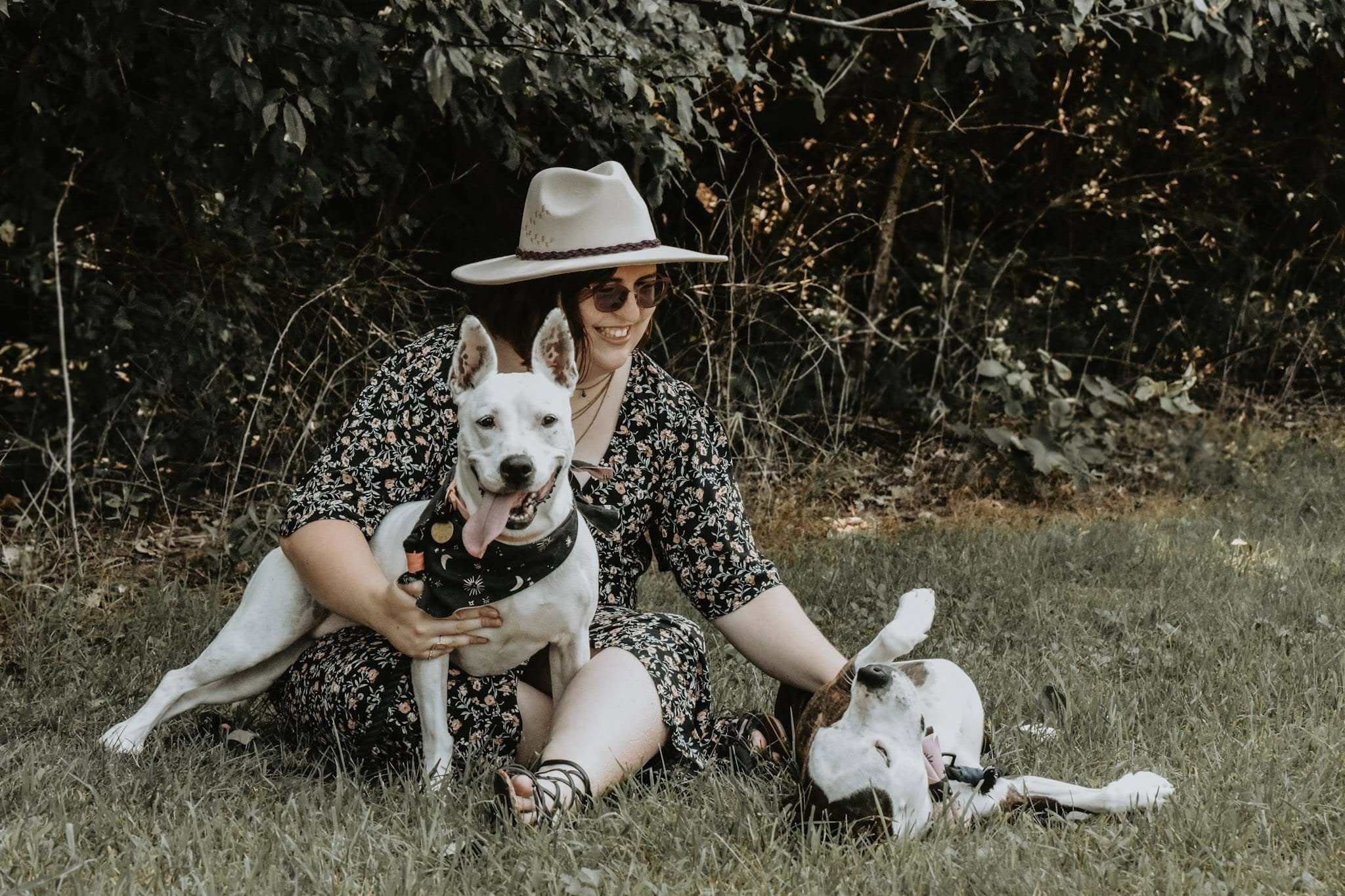 A woman sitting on grass outside, wearing a floral dress, sunglasses, a light-colored wide-brimmed hat, and smiling. She is holding a white dog with black spots and a bandana, and there are two other dogs lying at her feet. The background features dark foliage.