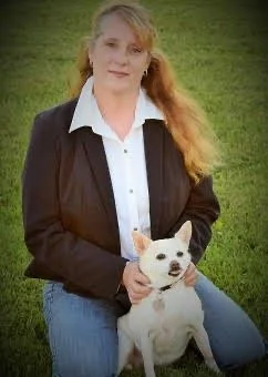 A young woman with blonde hair in a ponytail, wearing a black blazer and white shirt, standing outdoors on green grass, holding a small white dog with pointy ears.