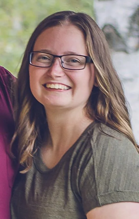 A young woman with glasses, long brown hair, and a green t-shirt, smiling outdoors.
