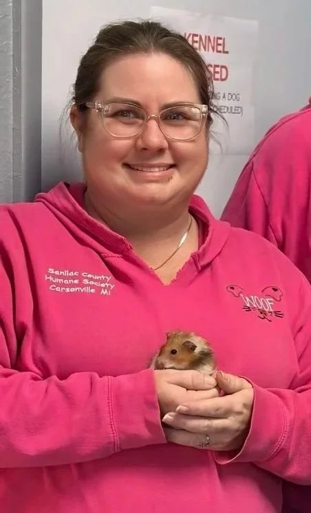A woman with glasses and brown hair pulled back, smiling, wearing a pink hoodie with text and graphic, holding a guinea pig.