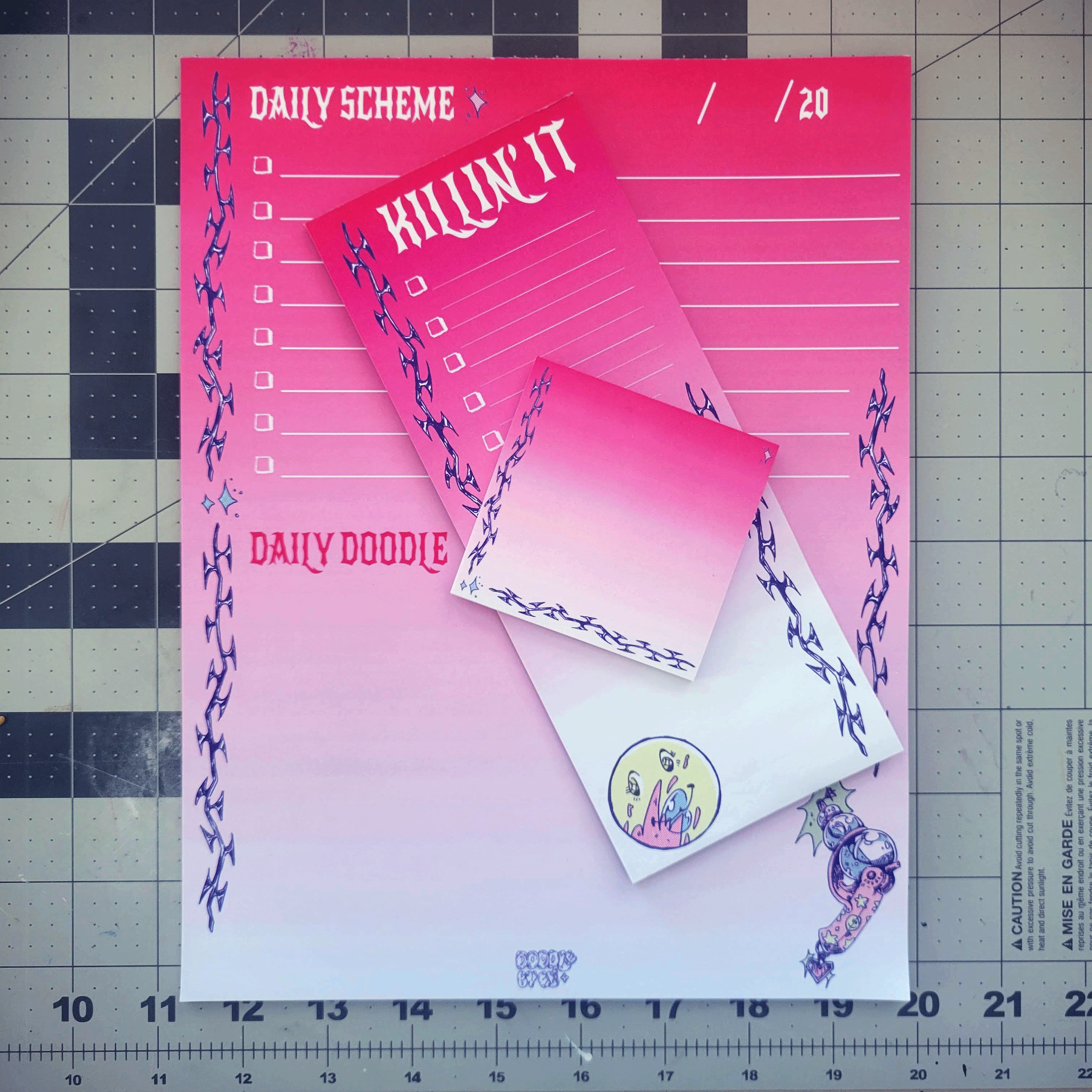 Pink and white stationery with designs and text, including checklists, labels, and cartoon graphics, placed on a cutting mat.