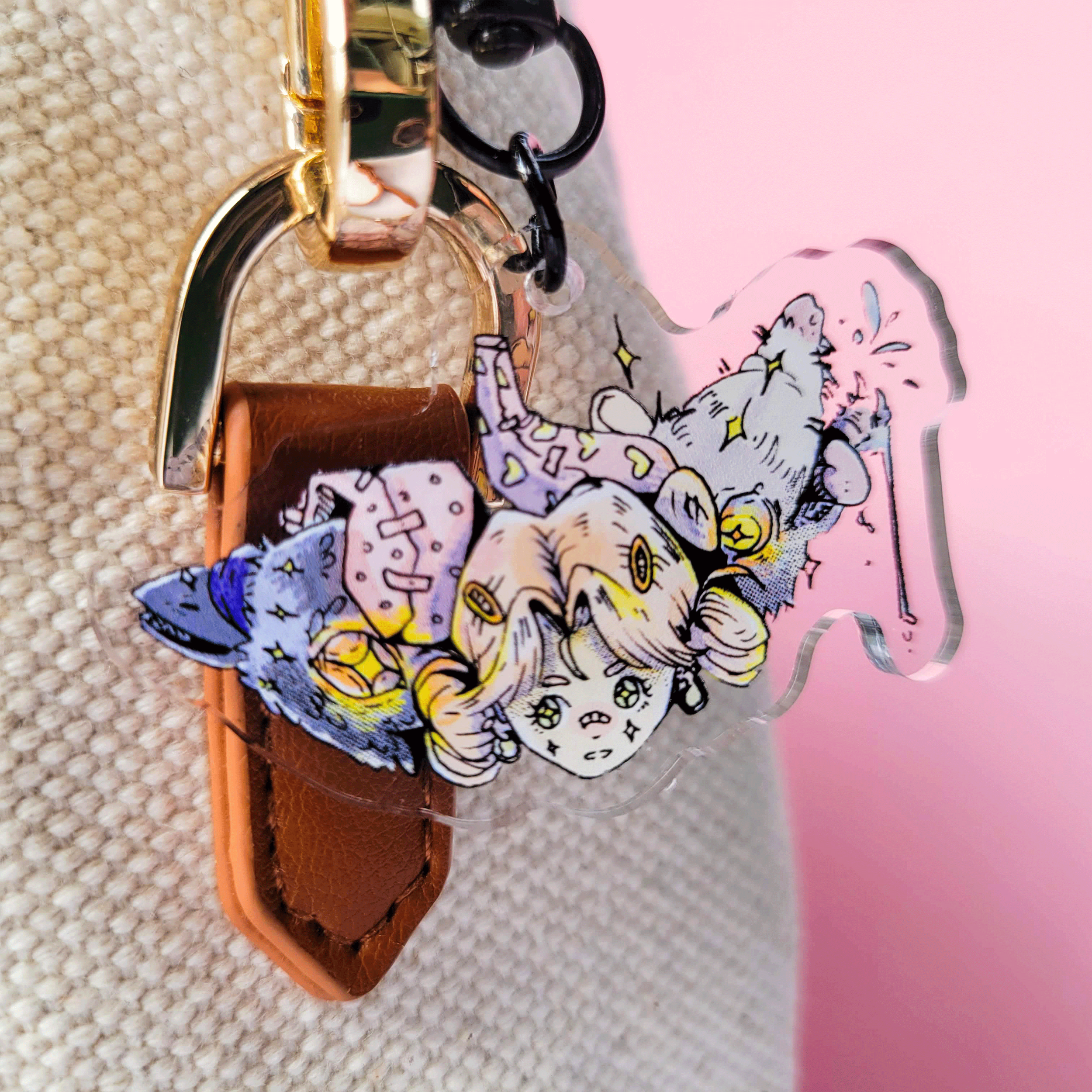 A keychain with a colorful cartoon character of a girl, along with two animal heads, a crow and a possum, attached to a beige canvas bag with a pink background.