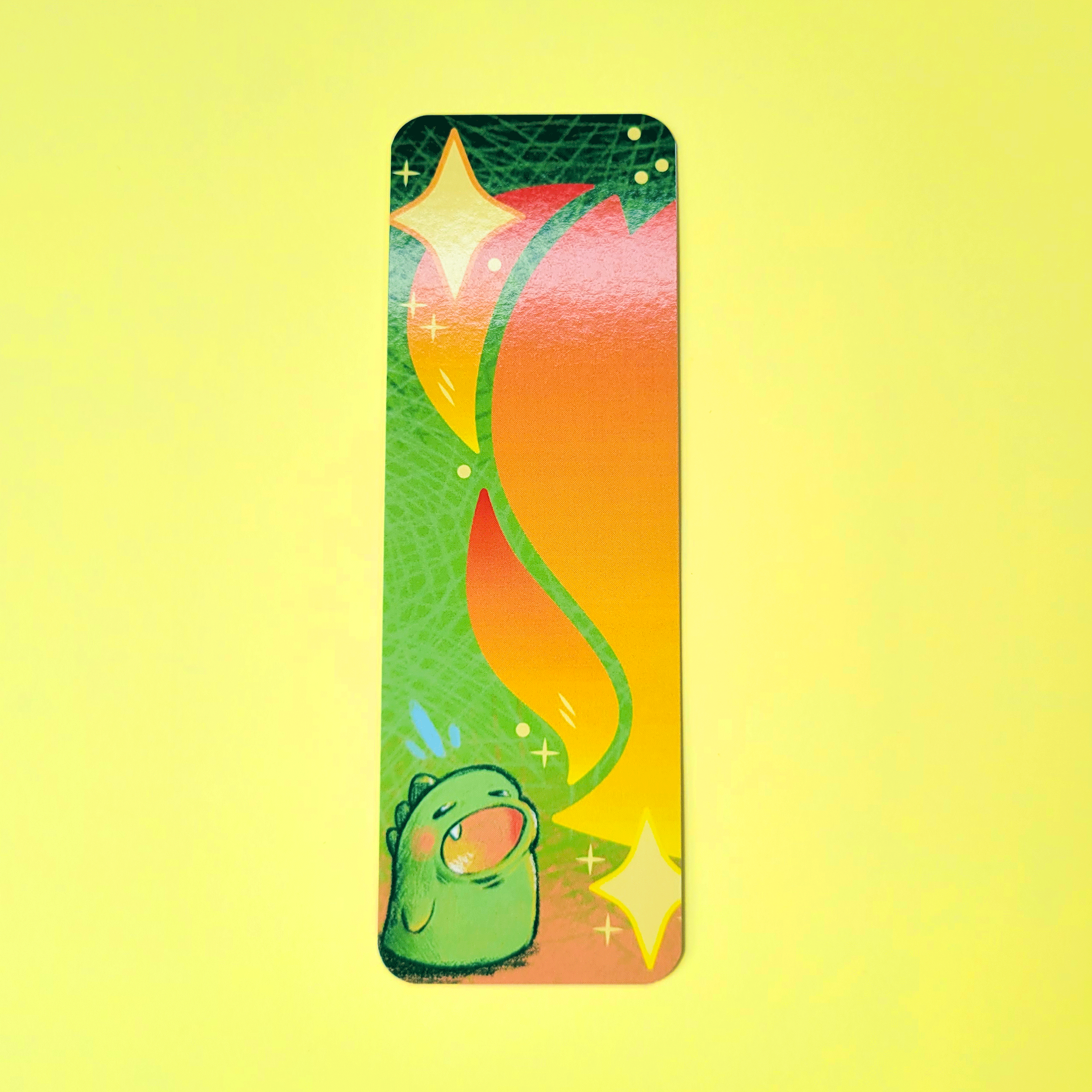 A colorful bookmark featuring a cute green dragon blowing fire. He is surrounded by swirling orange and yellow flames and sparkling stars on a vibrant background.