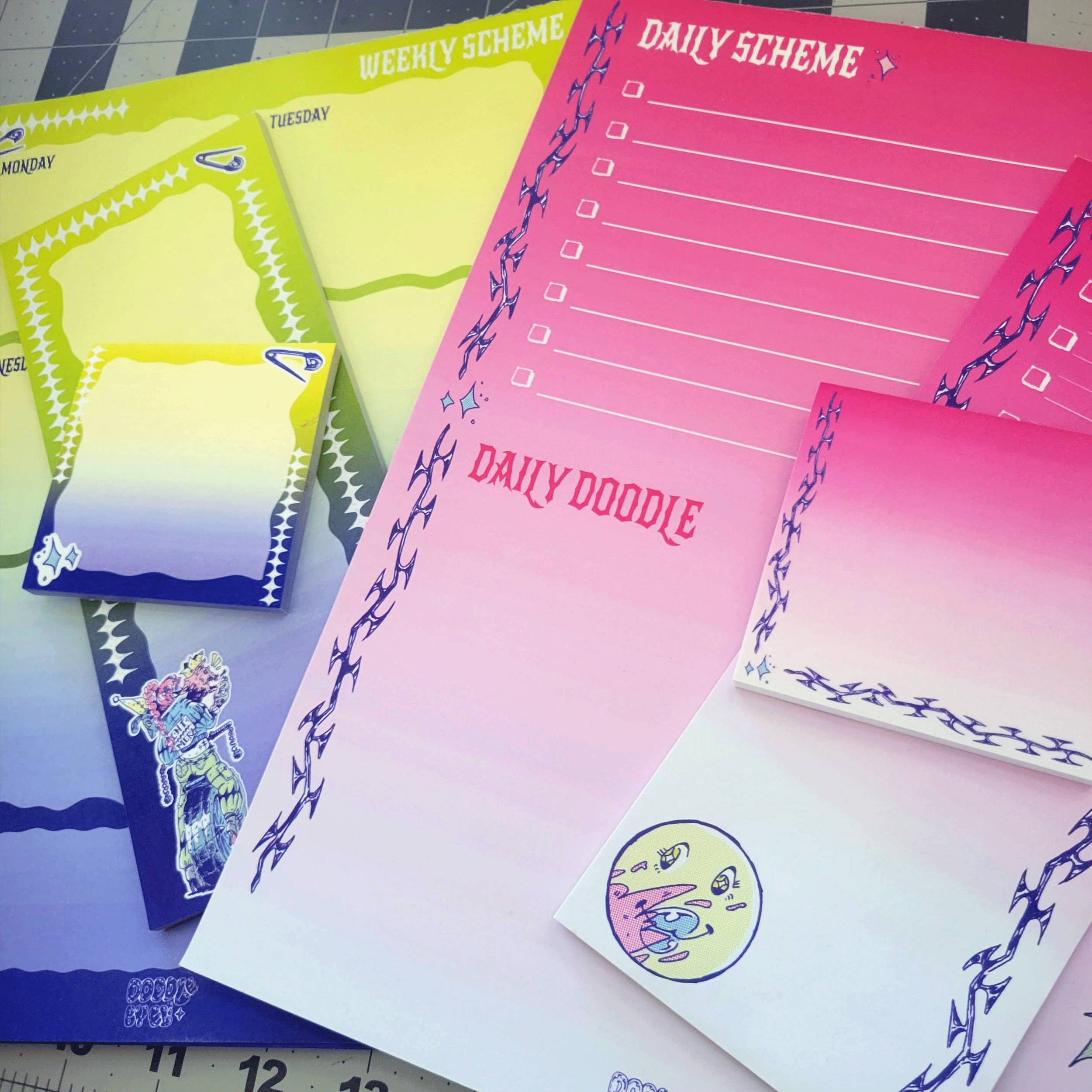 Colorful stationery with themes labeled "Weekly Scheme," "Daily Scheme," "Daily Doodle," and decorative borders, some featuring a smiling face and a girl on a punk-styled bicycle with a squirt gun and two animal heads coming out of her shoulders.