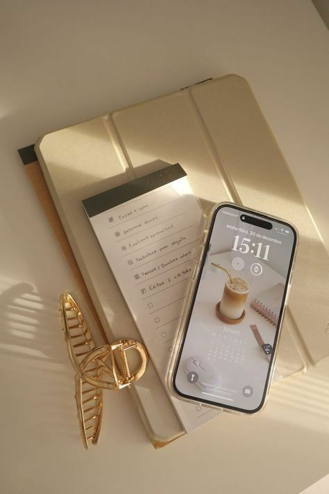 A beige notebook with a partially visible handwritten to-do list, a gold hairclip, and a smartphone displaying a coffee-themed wallpaper.