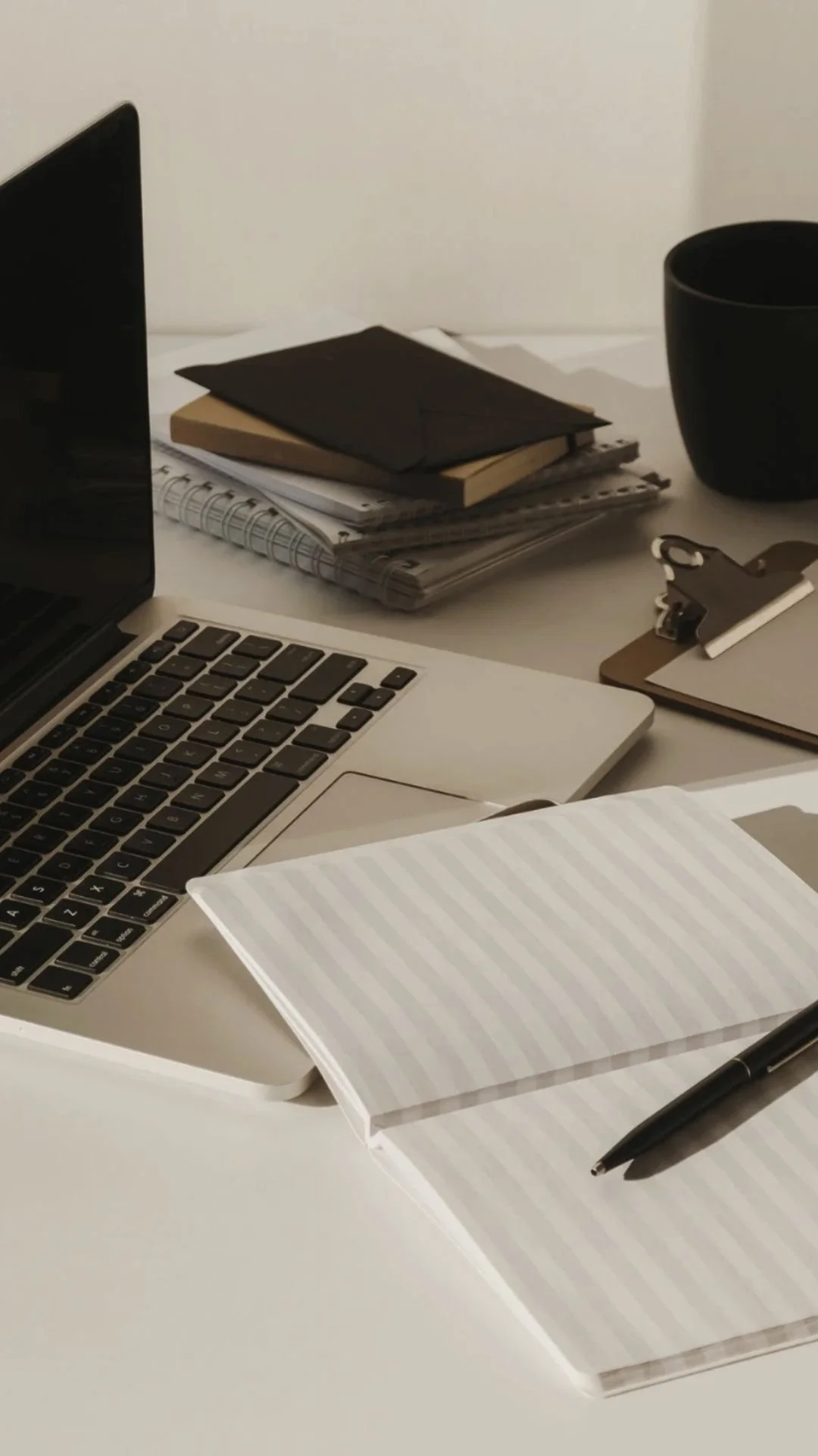 A workspace with a laptop, notebooks, a clipboard, a pen, and a black mug.