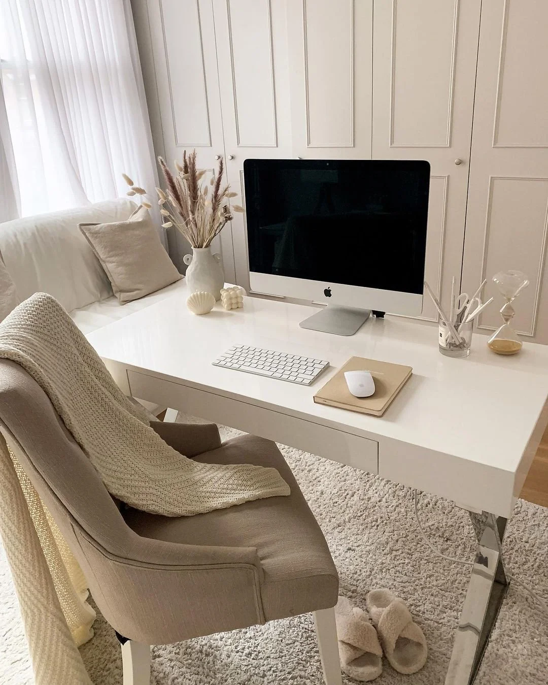 A cozy home office with a white desk, computer, keyboard, mouse, and beige chair with a knitted blanket. Decor includes a vase with dried flowers, candles, and decorative objects, all in neutral tones.