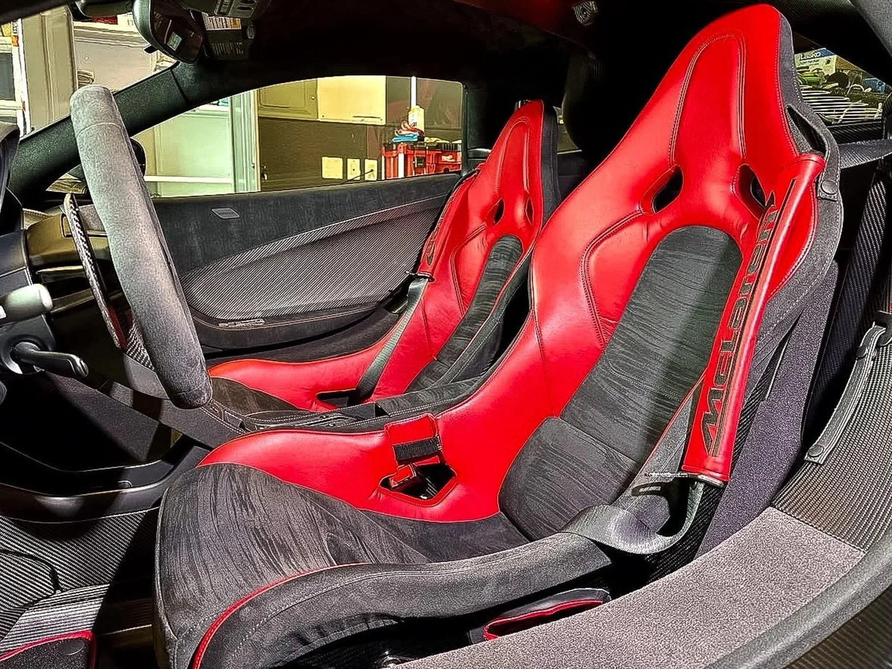 The interior of a McLaren car showing two racing seats, a black and red design. The seats are positioned side by side with a sleek, modern dashboard visible.