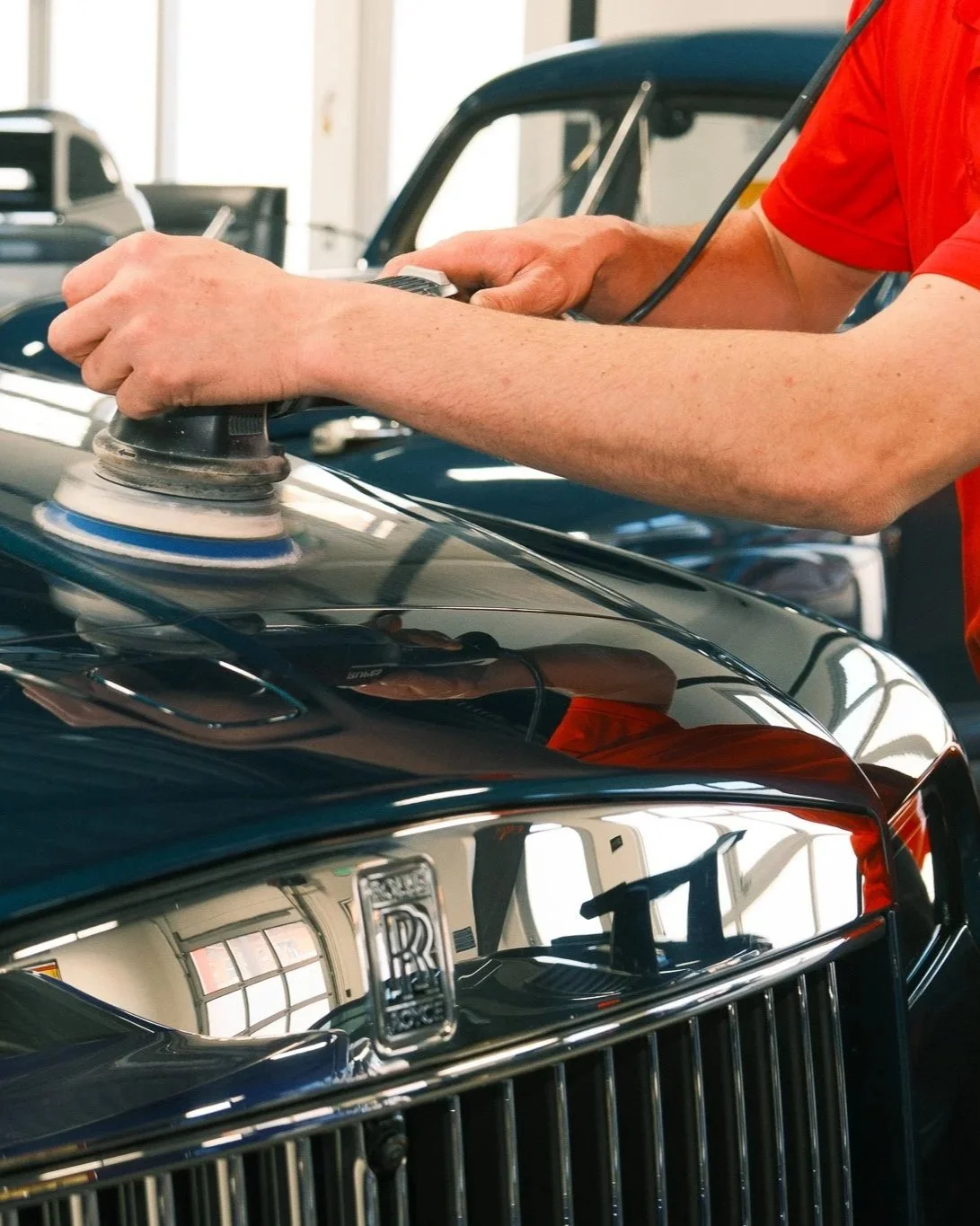 Person polishing the hood of a Rolls Royce with a buffer in a garage.