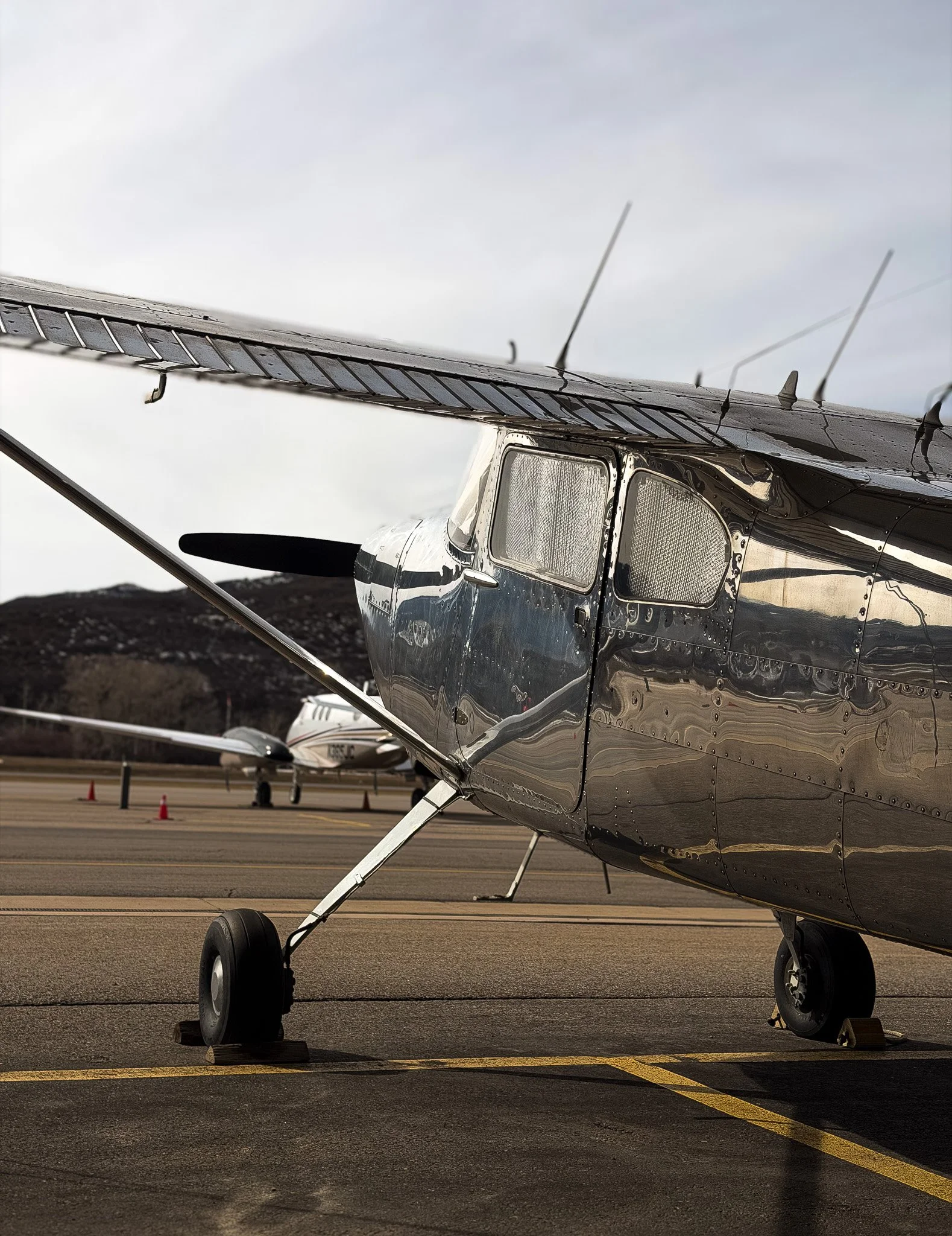 A shiny, metallic aircraft parked on the tarmac of an airport with other planes in the background under a cloudy sky. Aspen Mountain in the background at ASE airport